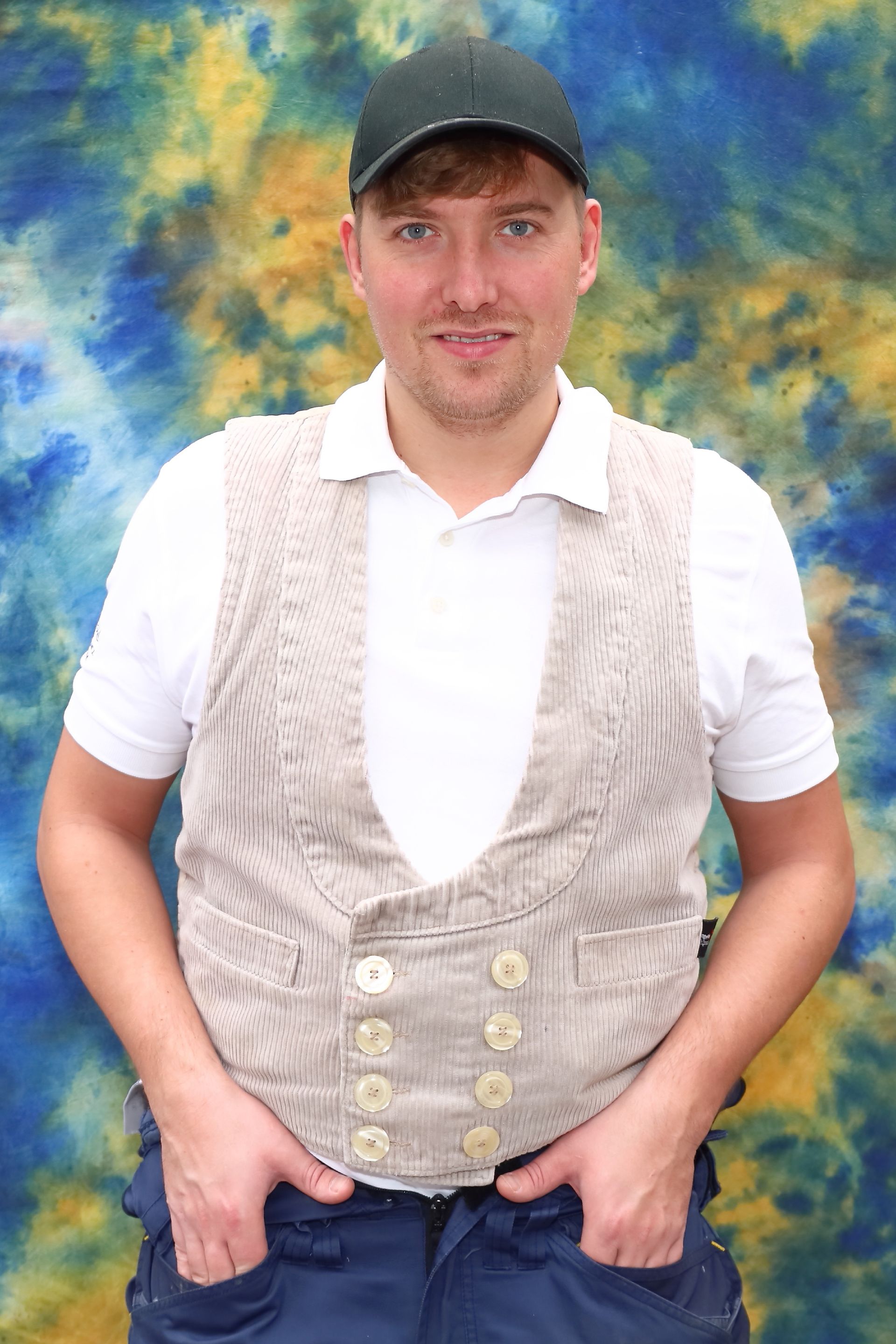 Mann in weißem Hemd, beiger Weste und dunkler Mütze, Hände in den Taschen. Blau-gelber Batik-Hintergrund.