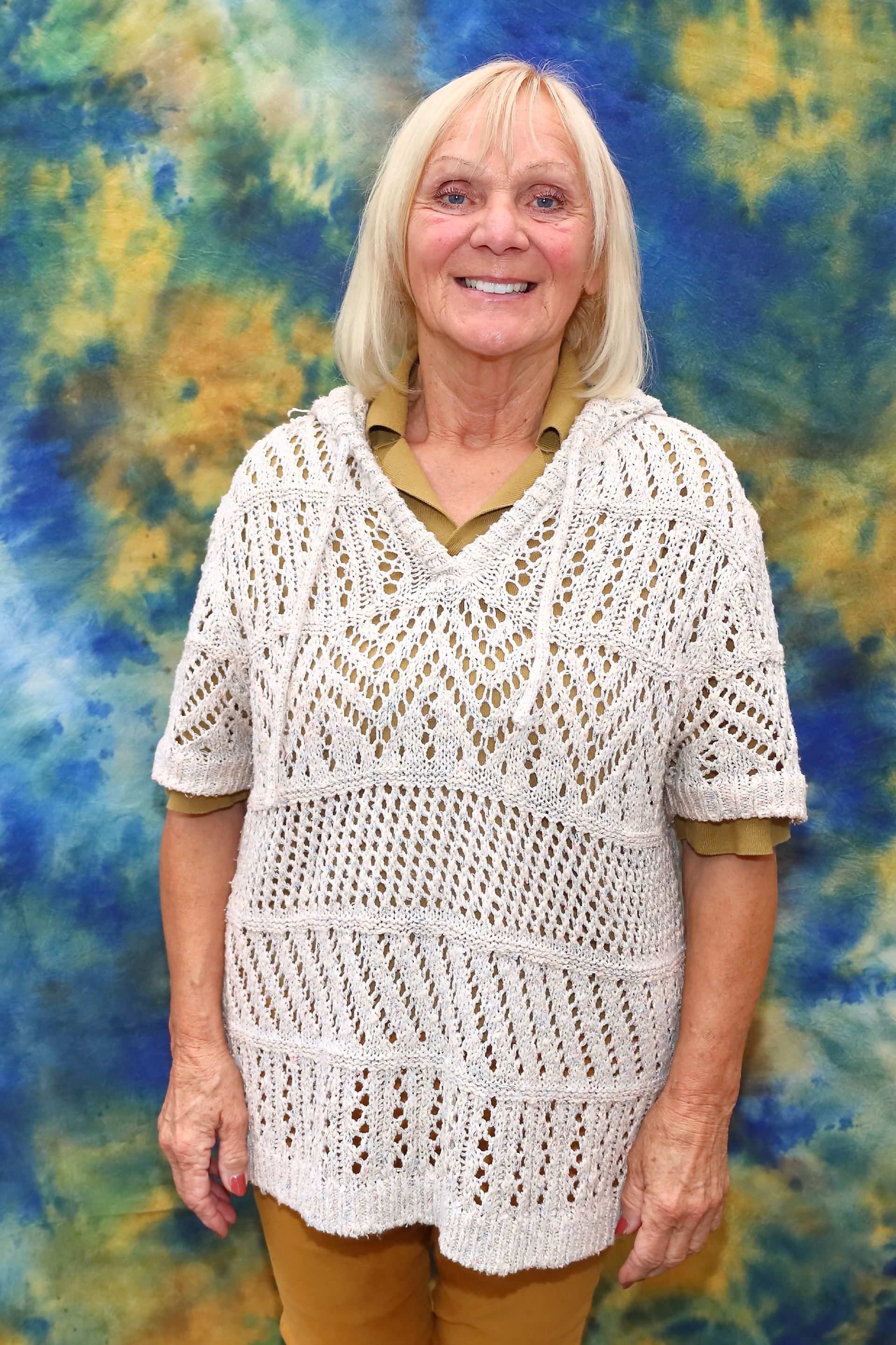 Eine lächelnde Frau trägt einen weißen Strickpullover über einer senffarbenen Bluse und steht vor einem blau-gelben Hintergrund.