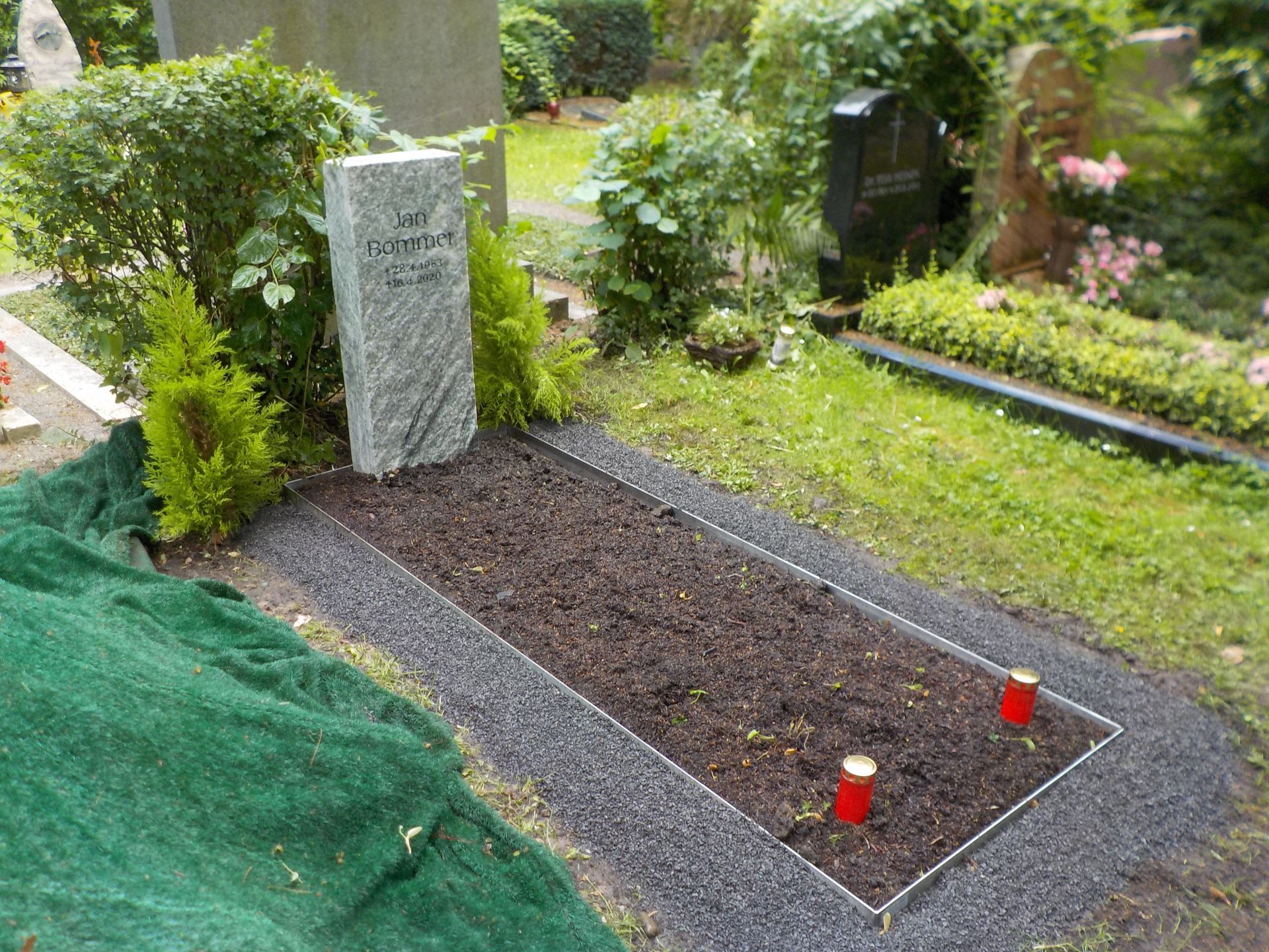 Grab mit grauem Grabstein, dunkler Erde und schwarzem Rand; zwei rote Kerzen; grünes Laubwerk.