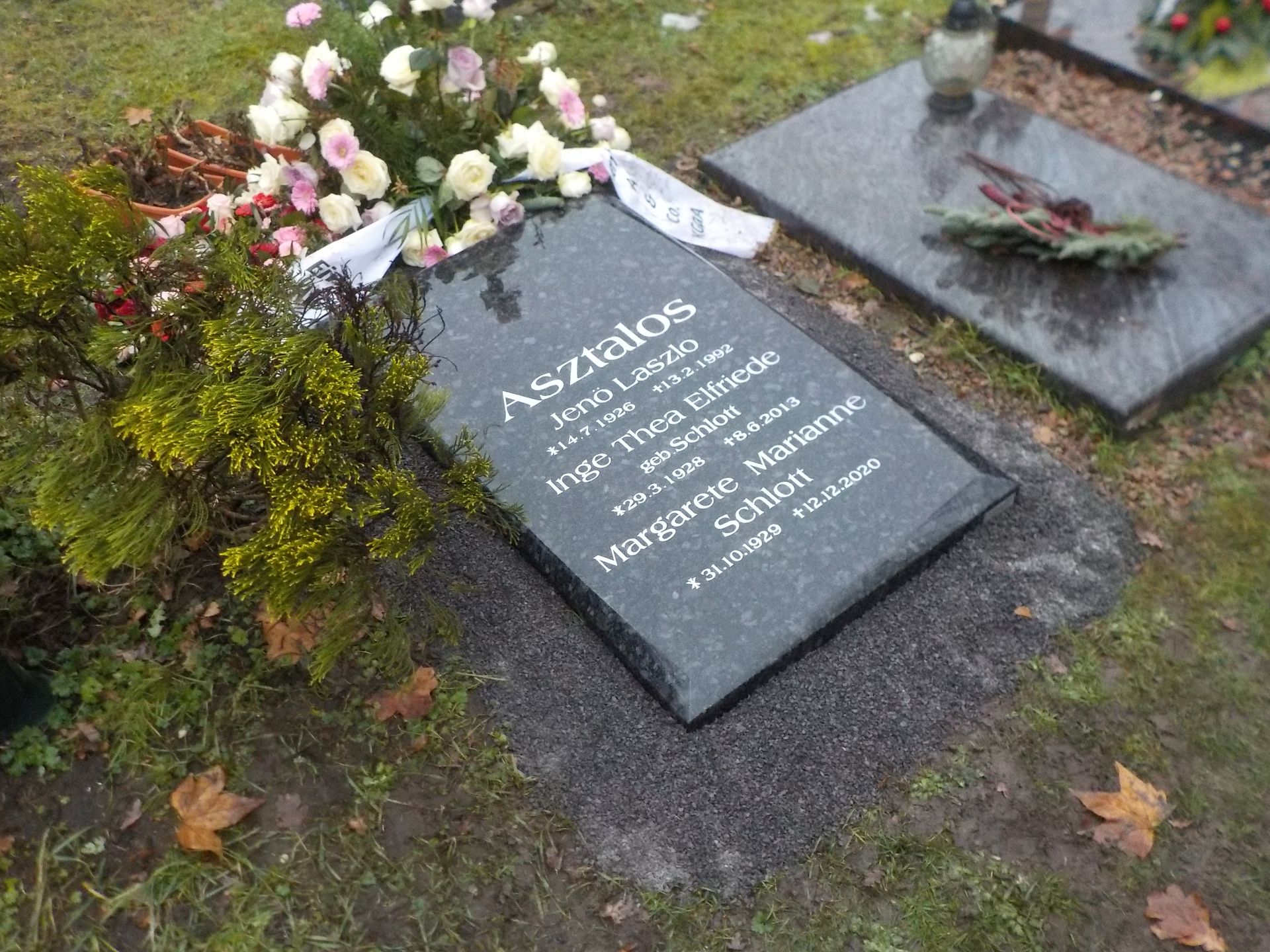 Grabstein mit Blumen; schwarzer Stein mit eingravierten Namen: Asztalos, Irén usw. Grauer Kies, bewölkter Tag.