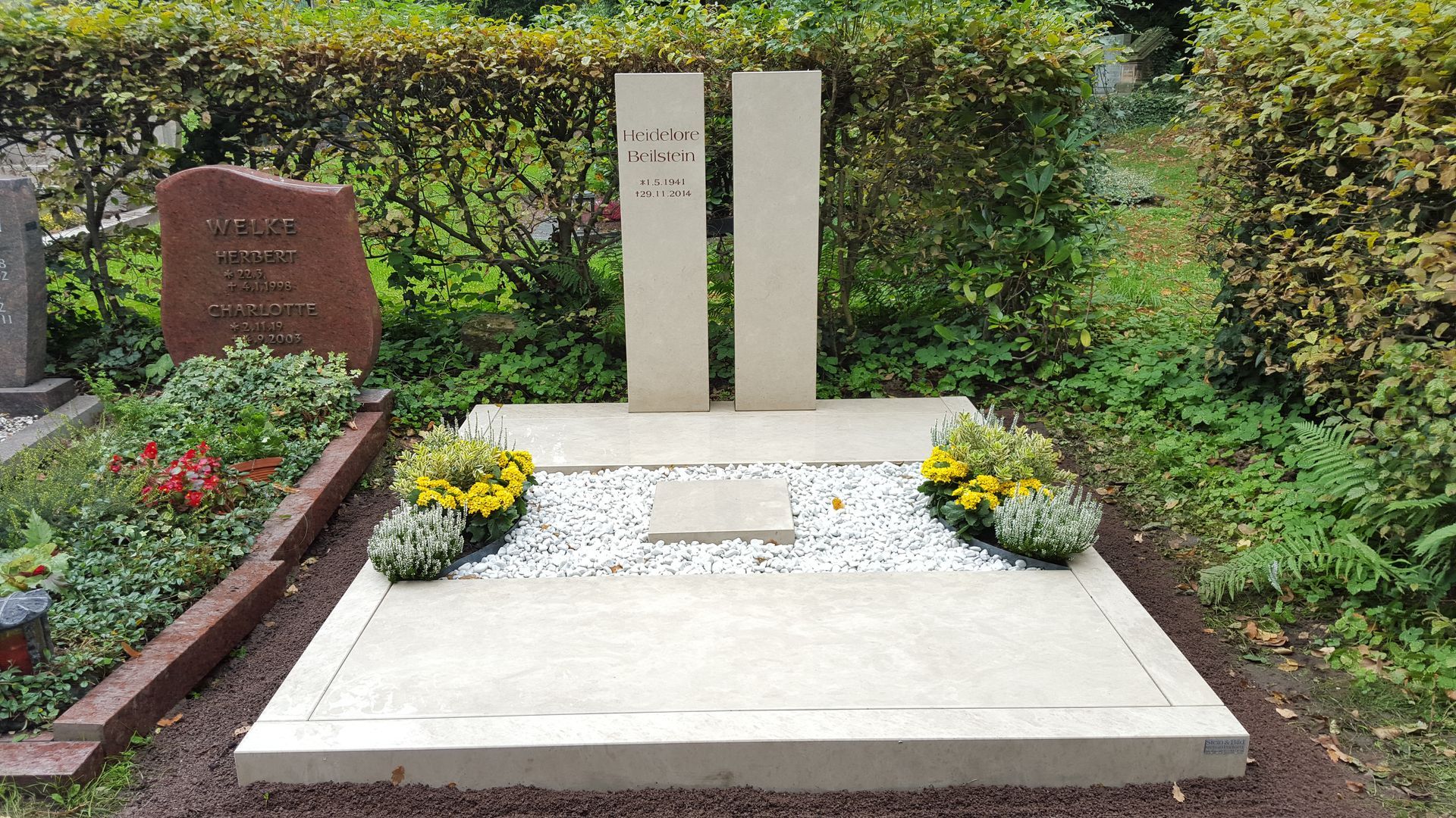 Grabstein mit zwei aufrecht stehenden, hellen Steinsäulen, flachem Sockel und weißem Kies, verziert mit Grün und Blumen.