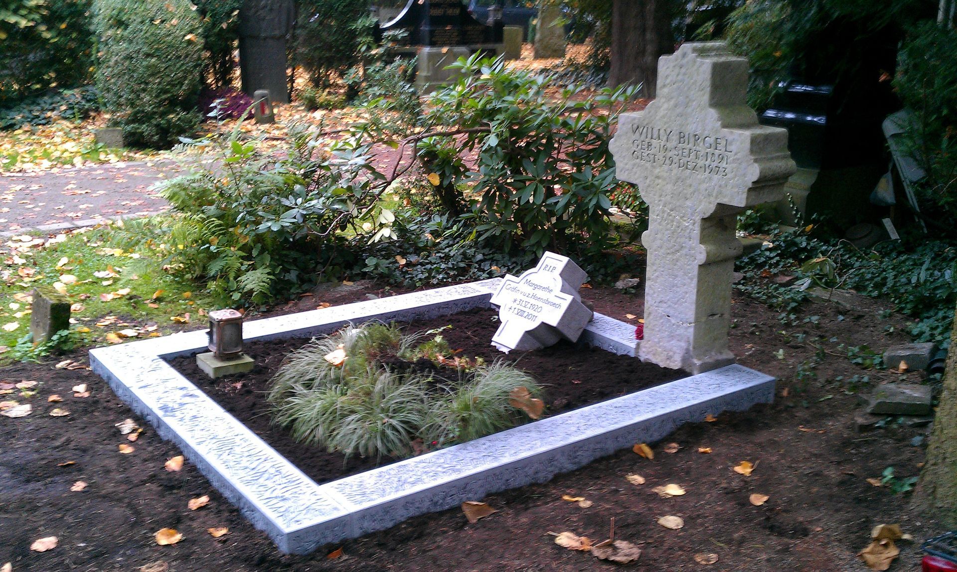 Ein Steingrab mit einem keltischen Kreuz, umgeben von einer Einfassung und kleinen Pflanzen, in einer Friedhofsanlage.
