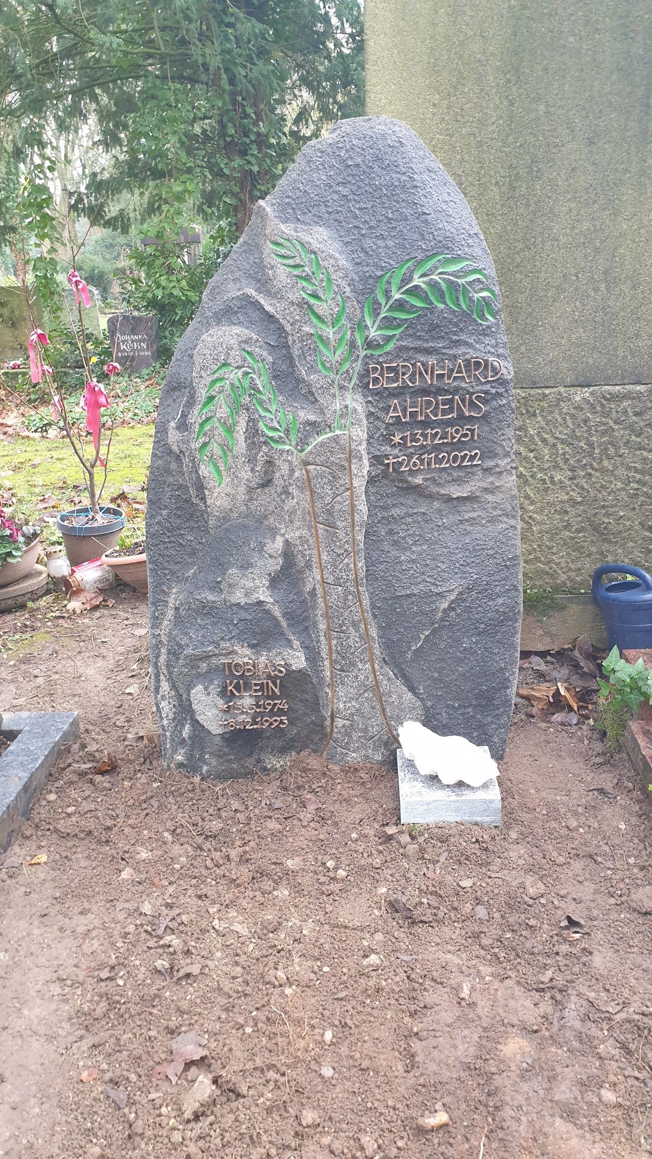 Grabstein im Garten. Hoher, runder Stein mit Inschriften. Grüne Blumenverzierungen. Braune Erde ringsum.