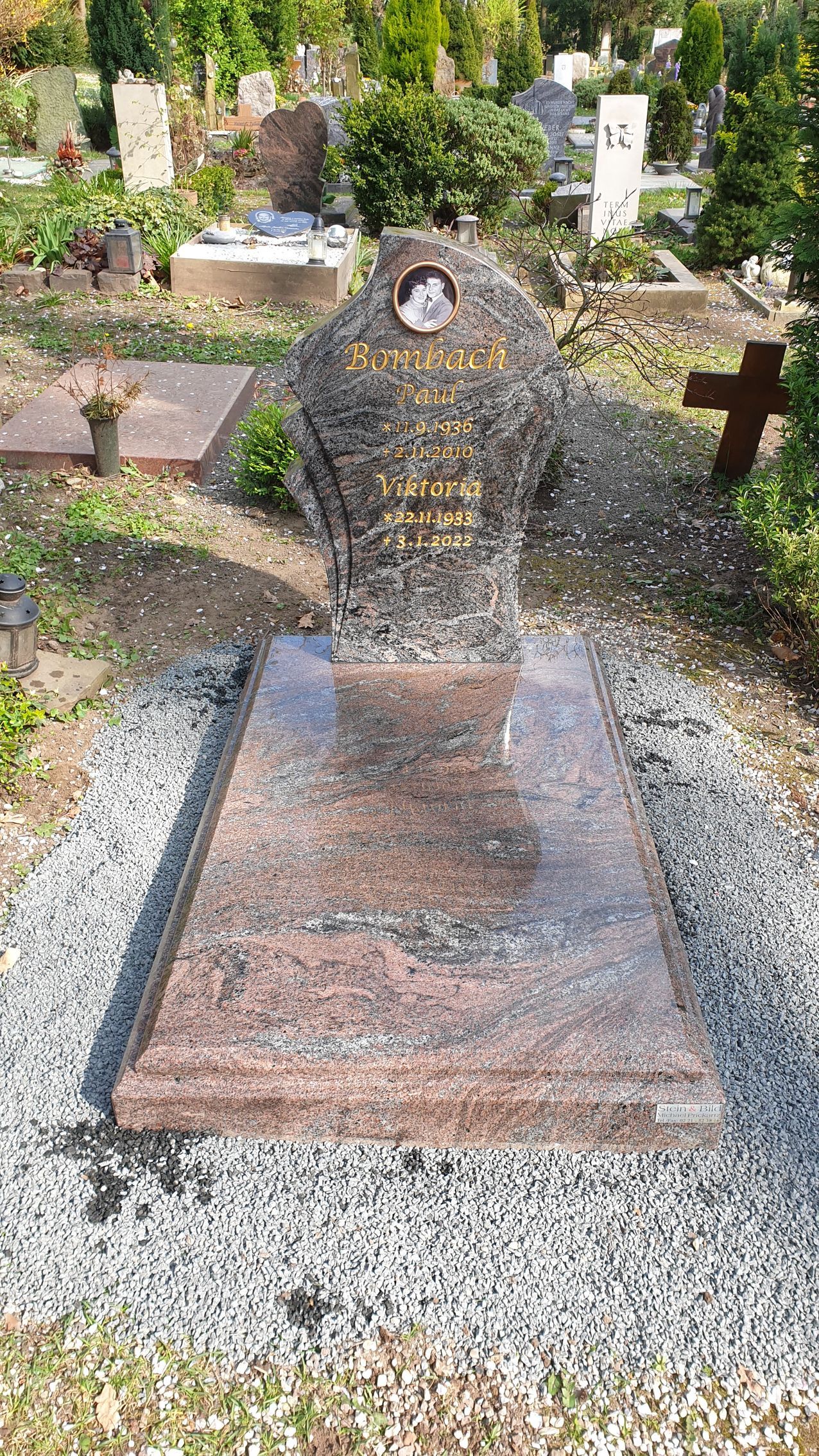 Grab auf einem Friedhof. Ein steinerner Grabstein mit einem Porträt steht auf einer großen, polierten, rotbraunen Granitplatte.