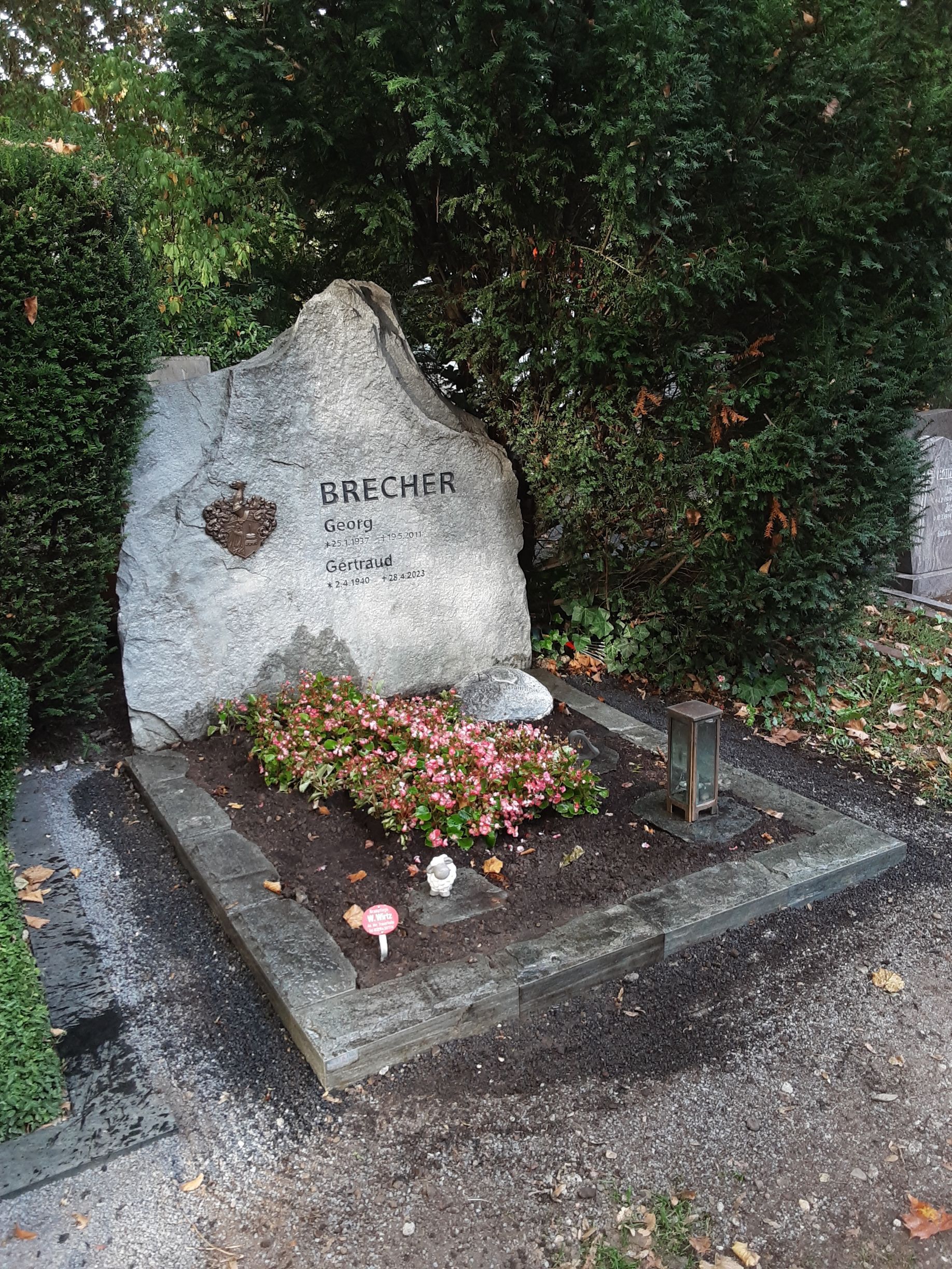 Grab mit einem großen, strukturierten Steingrabstein. Blumen und eine kleine Statue zieren das Grab. Im Hintergrund ist grünes Laubwerk zu sehen.