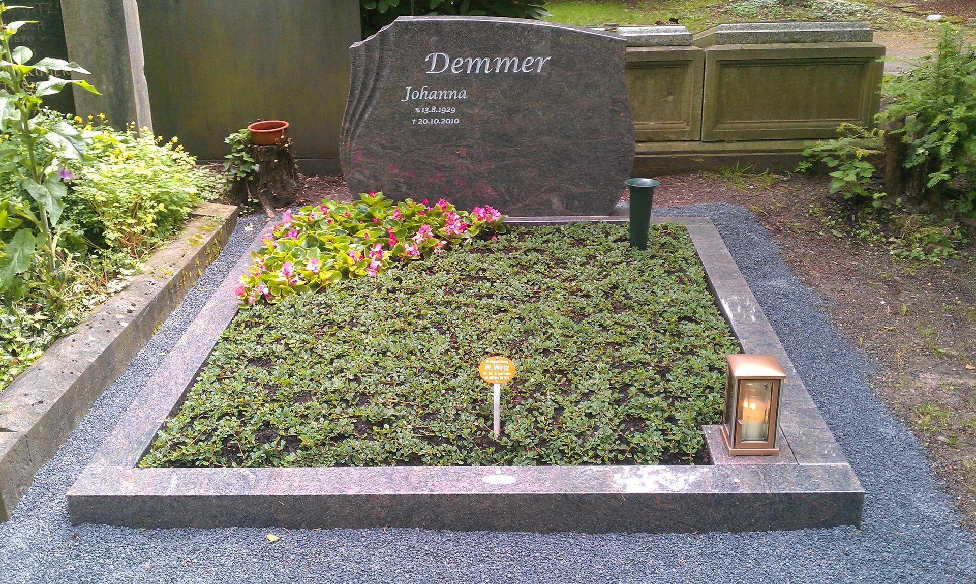 Grabstätte mit Grabstein, Blumen und Grünbewuchs. Dunkelgrauer Grabstein, hellgrauer Rand.