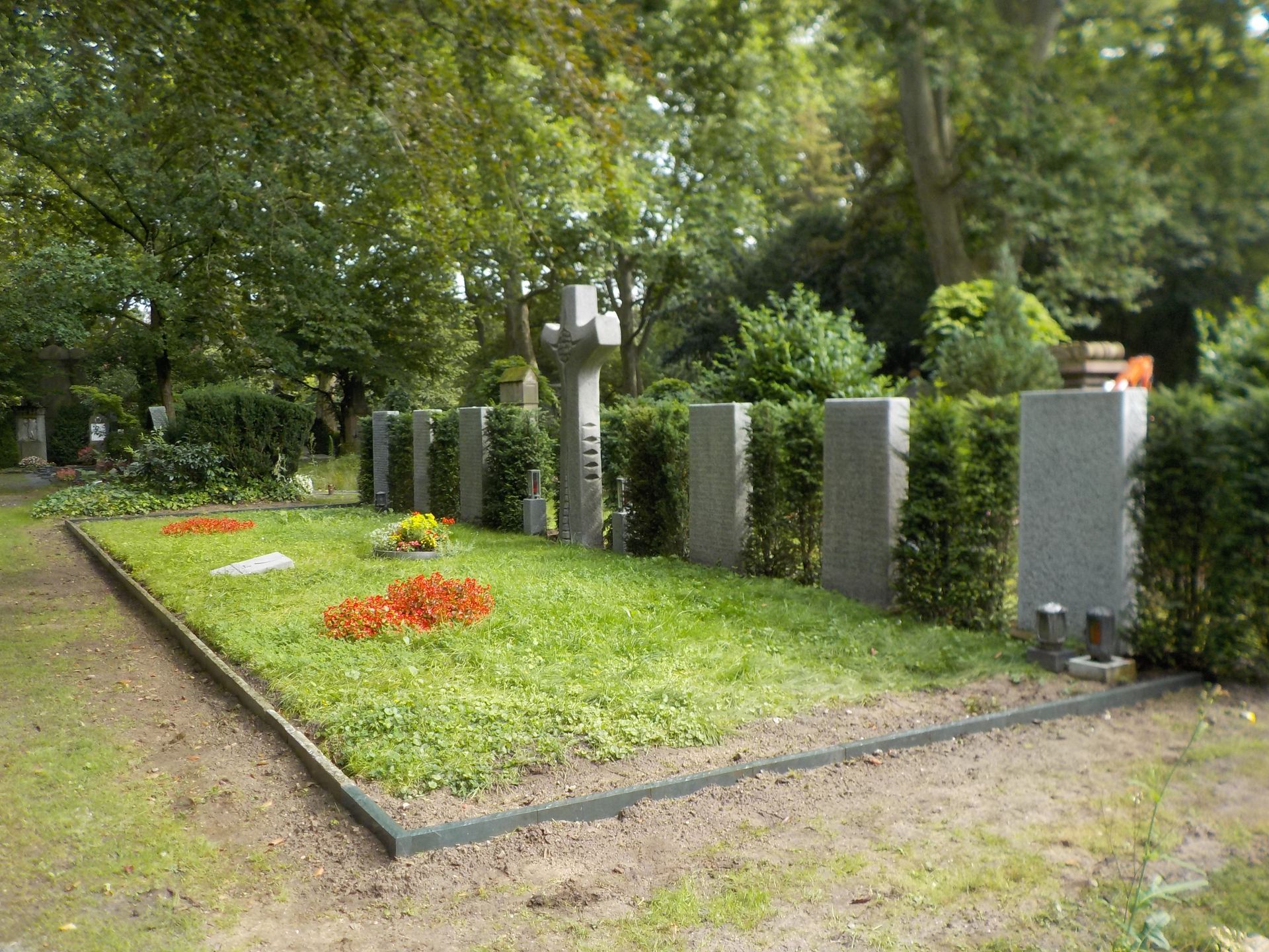 Grabstätte auf einem Friedhof mit rechteckigem Grundstück, grünem Gras, steinernen Grabsteinen und blühenden orangefarbenen und gelben Blumen.