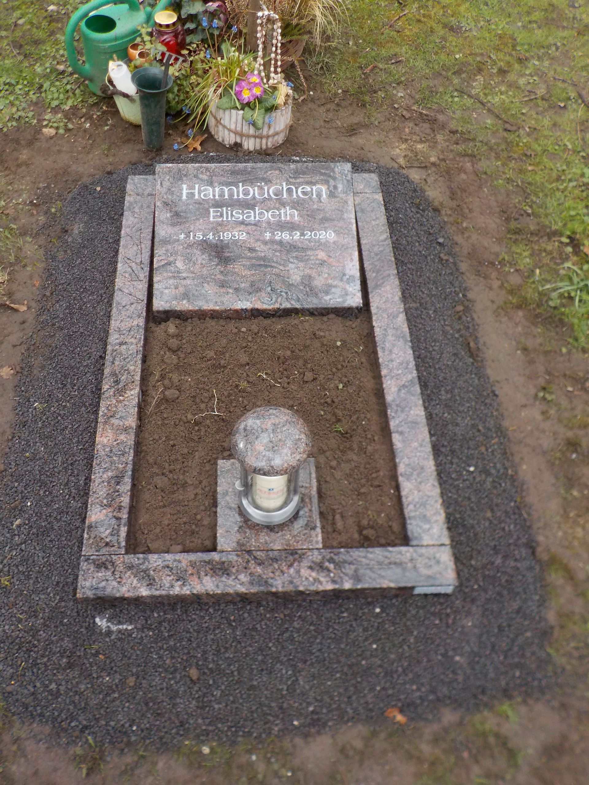 Grab mit Granitgrabstein und -einfassung, Erde und einer Laterne. Eine Gießkanne und Blumen liegen daneben.