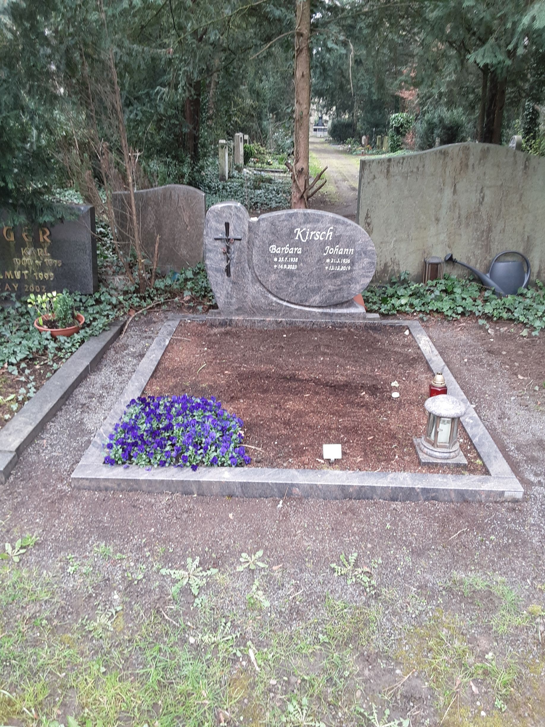 Grabstätte mit einem steinernen Grabstein, blauen Blumen und einer Gedenklaterne. Friedhofsumgebung.