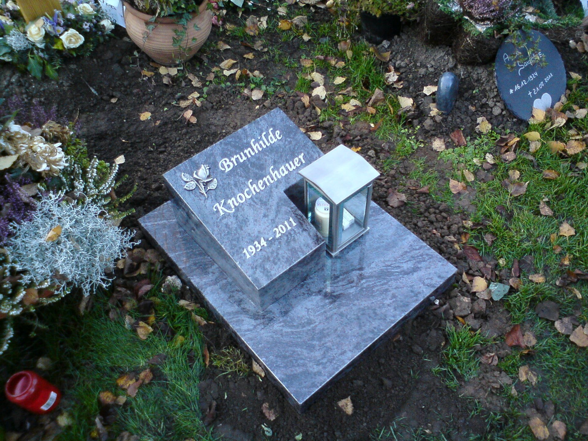 Grab mit grauem Granitgrabstein, Laterne und Blumenschmuck auf einer Rasenfläche, verstreutes Laub.