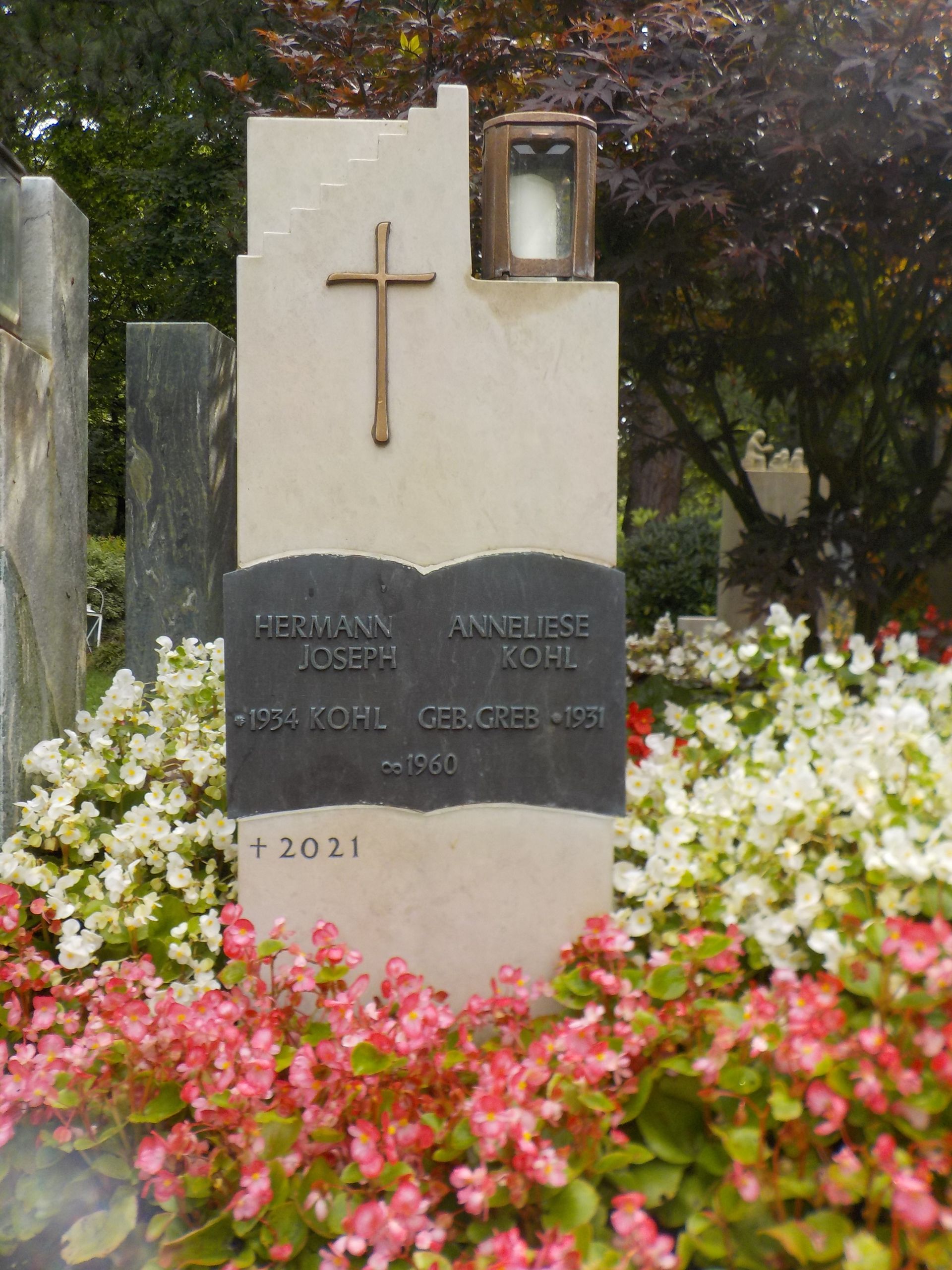 Grabstein mit Kreuz, Namen und Daten, umgeben von Blumen.