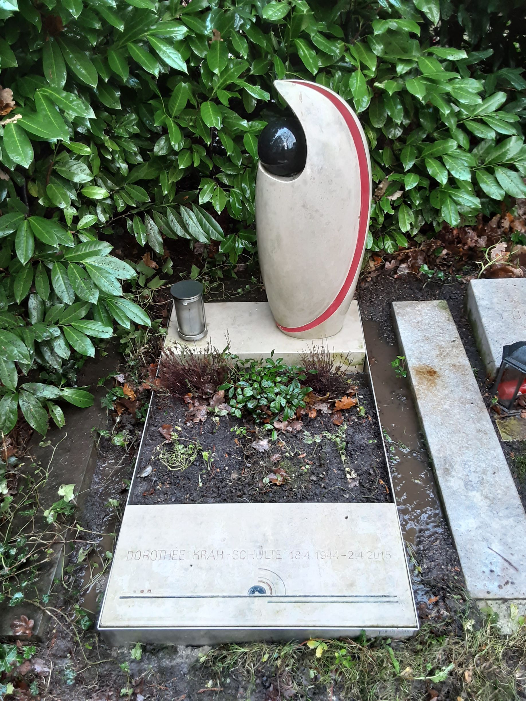 Grab mit moderner Skulptur, schwarzer Kugel, cremefarbenen und roten Akzenten, nahe grünem Laubwerk.