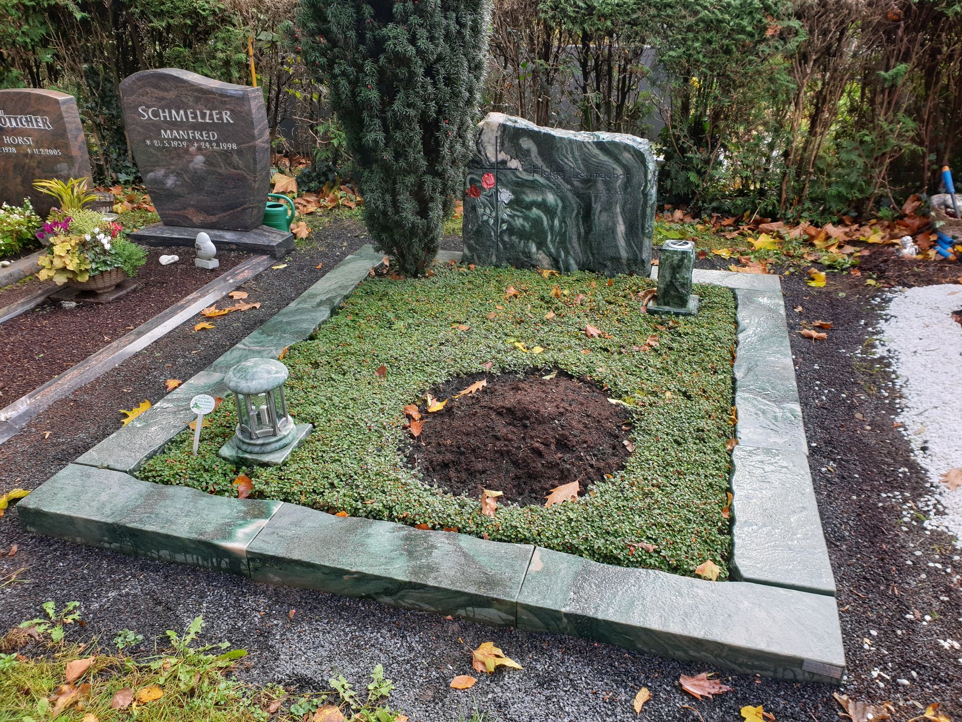 Grabstätte mit grünem Gras und einer dunklen, frisch ausgehobenen Stelle. Granitgrabstein, Laterne und umgebende Steine.