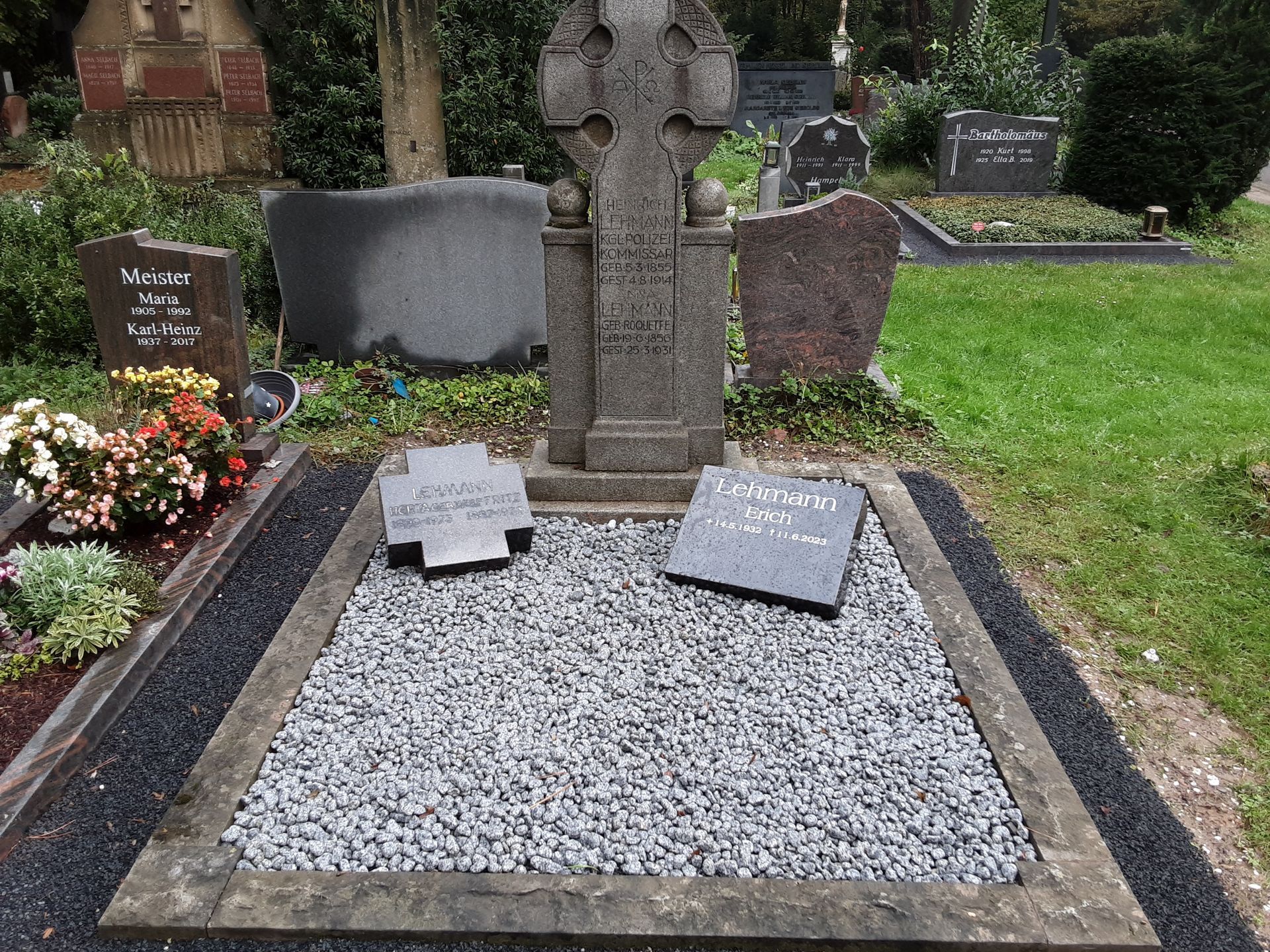 Grabstein auf einem Friedhof, bestehend aus einem hohen Kreuz, zwei flachen Markierungen und Kies.