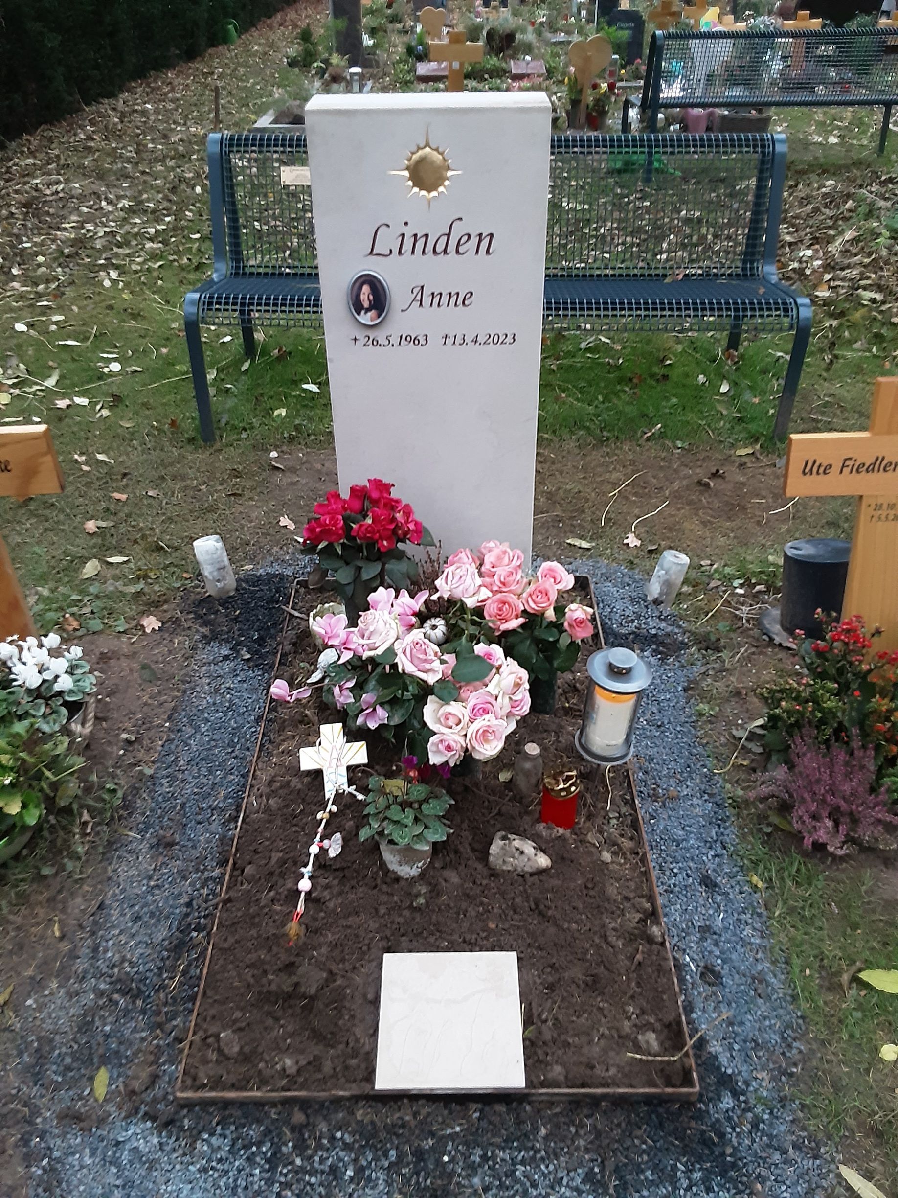 Grabstein mit Blumen auf einem Friedhof. Er trägt ein Foto, den Namen und die Lebensdaten. Rosa und rote Blumen schmücken das Grab.