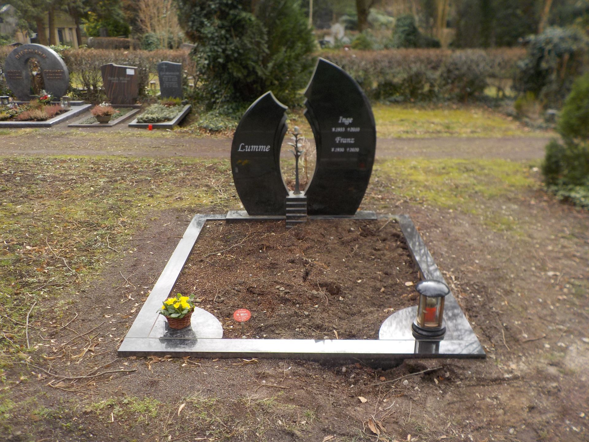 Schwarzer Granitgrabstein mit gespaltenem Muster; ein Grab mit Metallrand; Blumen und eine Laterne.