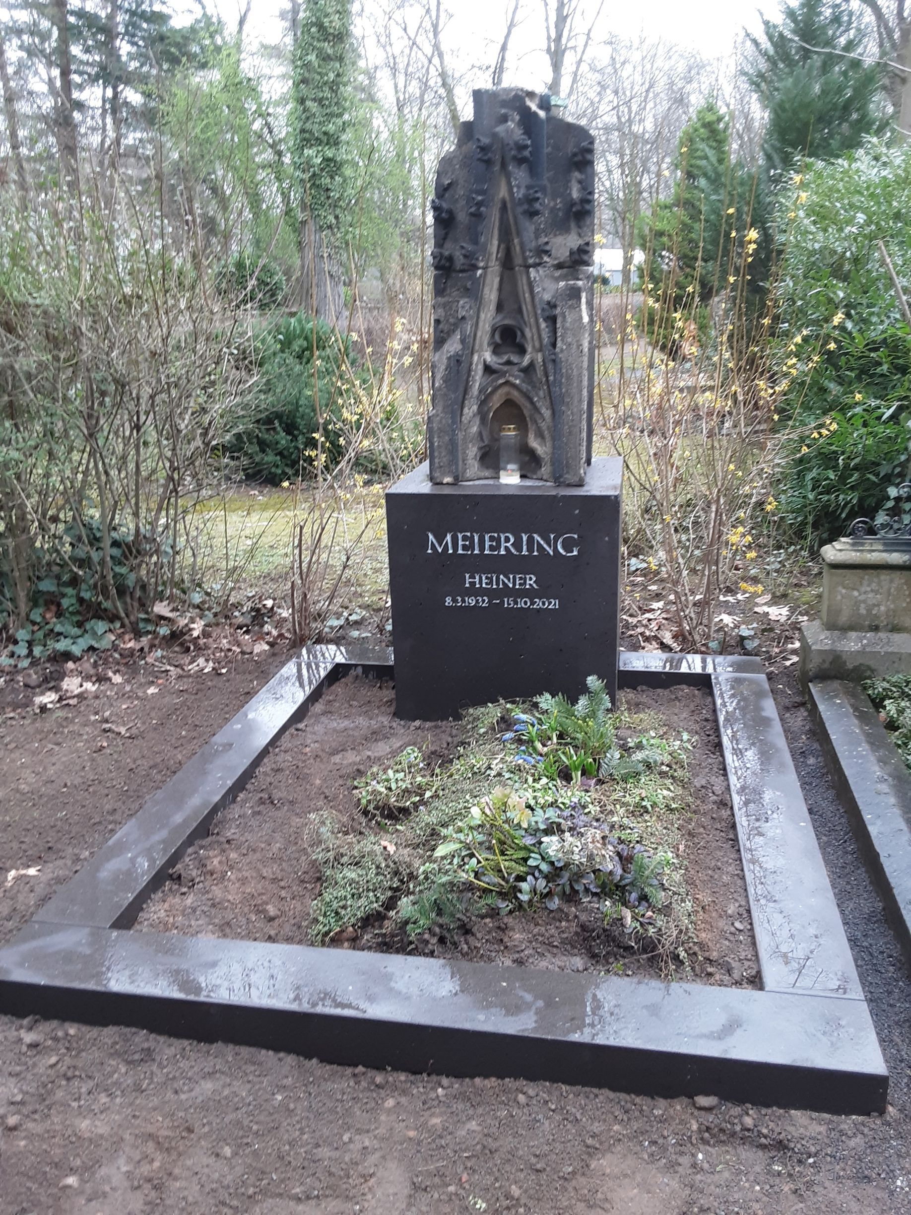 Grab mit einem dunklen Steinmonument und einer Namensinschrift. Umgeben von einer Einfassung und kleinen Pflanzen. Im Freien.