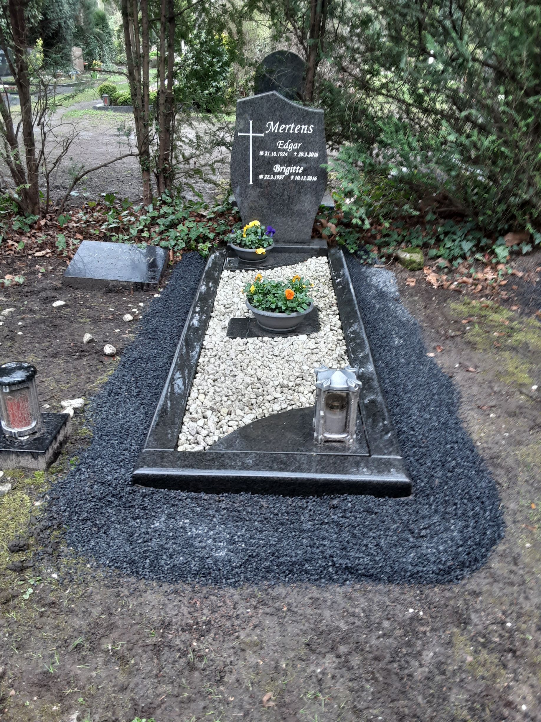 Grabstätte mit dunklem Grabstein, Blumen und einem Bett aus schwarzen und weißen Steinen. Umgeben von dunklem Kies.