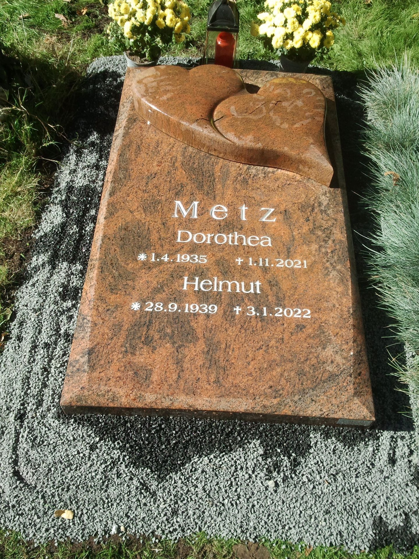 Grabstein für die Familie Metz, Granit, eingravierte Namen und Daten. Rotbrauner Stein. Blumen im Hintergrund.