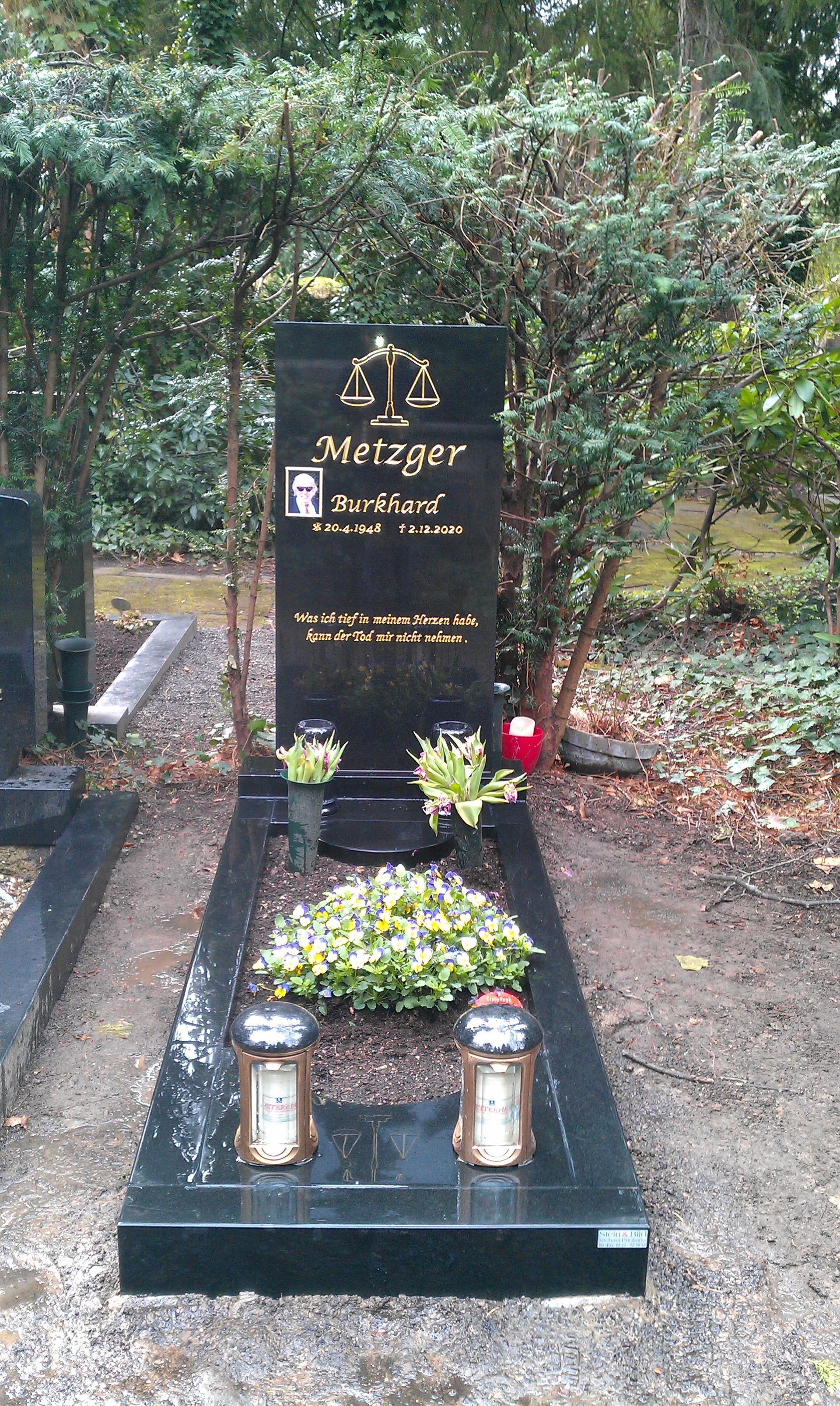 Schwarzer Granitgrabstein mit Sockel auf einem Friedhof, umgeben von einem Kranz, Kerzen und Bäumen.