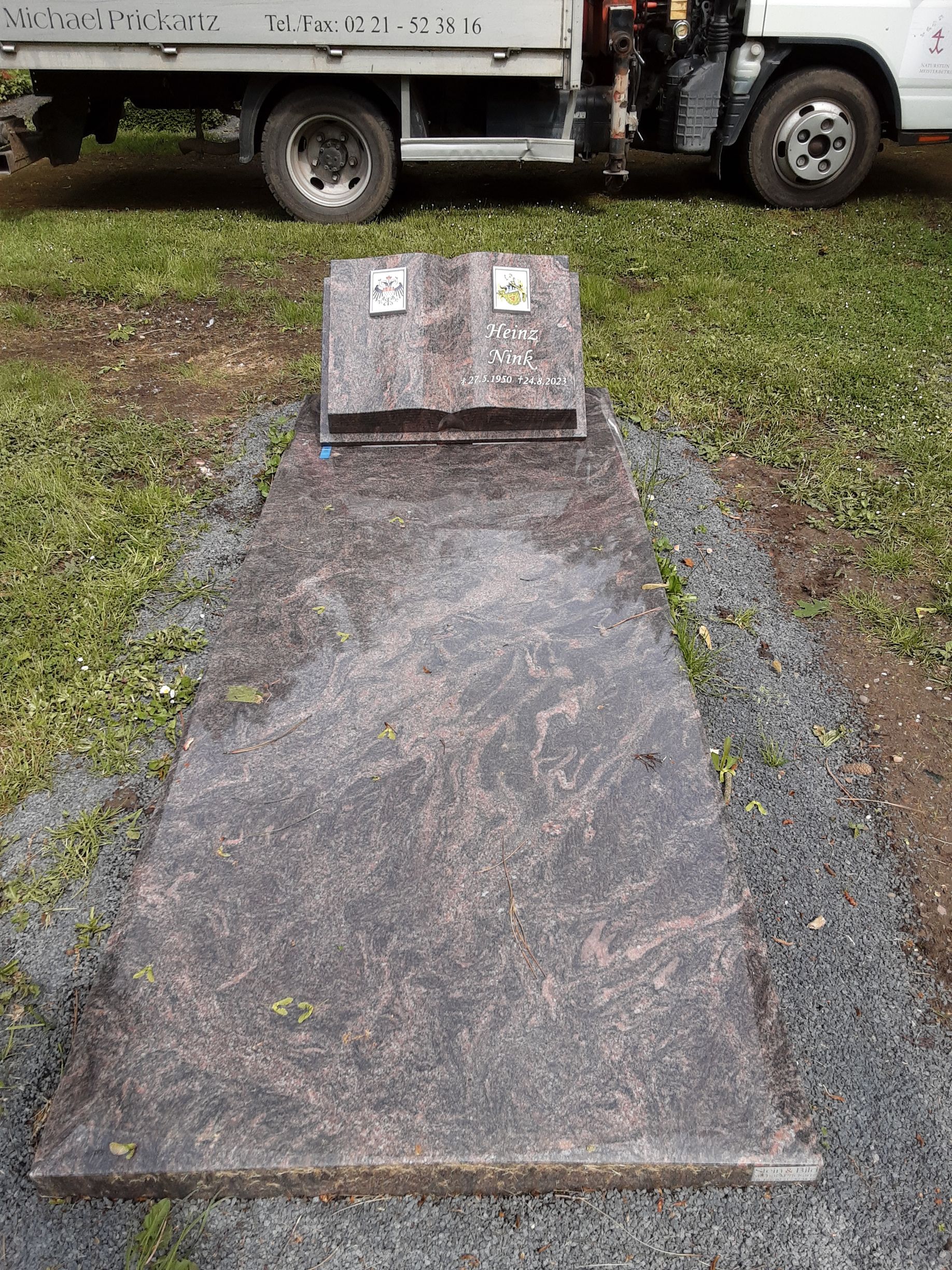 Grabstein aus Granit mit rechteckigem Sockel und erhöhtem Grabsteinteil; Fahrzeug im Hintergrund.