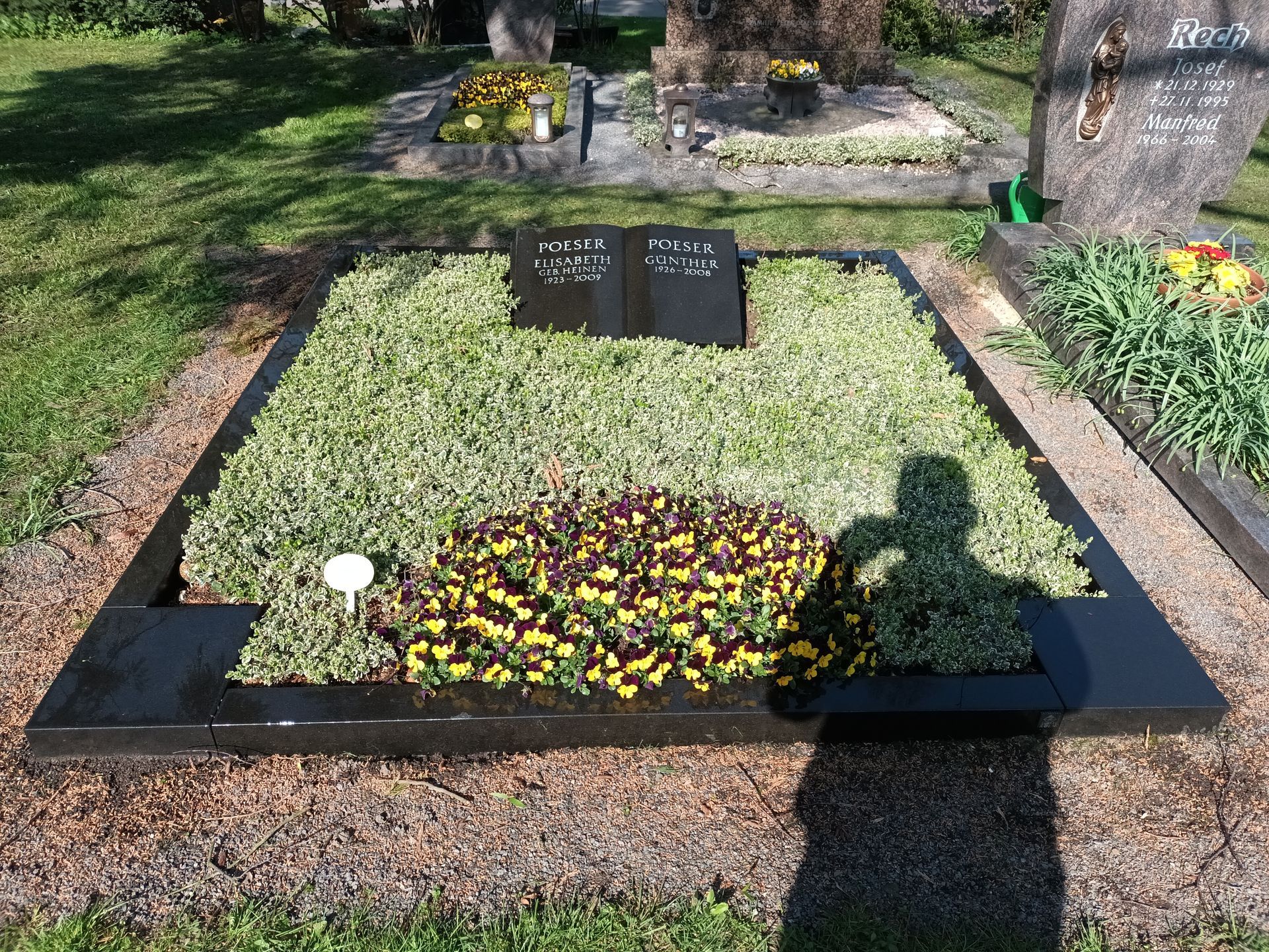 Grab mit schwarzem Grabstein, umrandet von schwarzem Grün und bepflanzt mit Pflanzen und Blumen. Ein Schatten fällt darauf.
