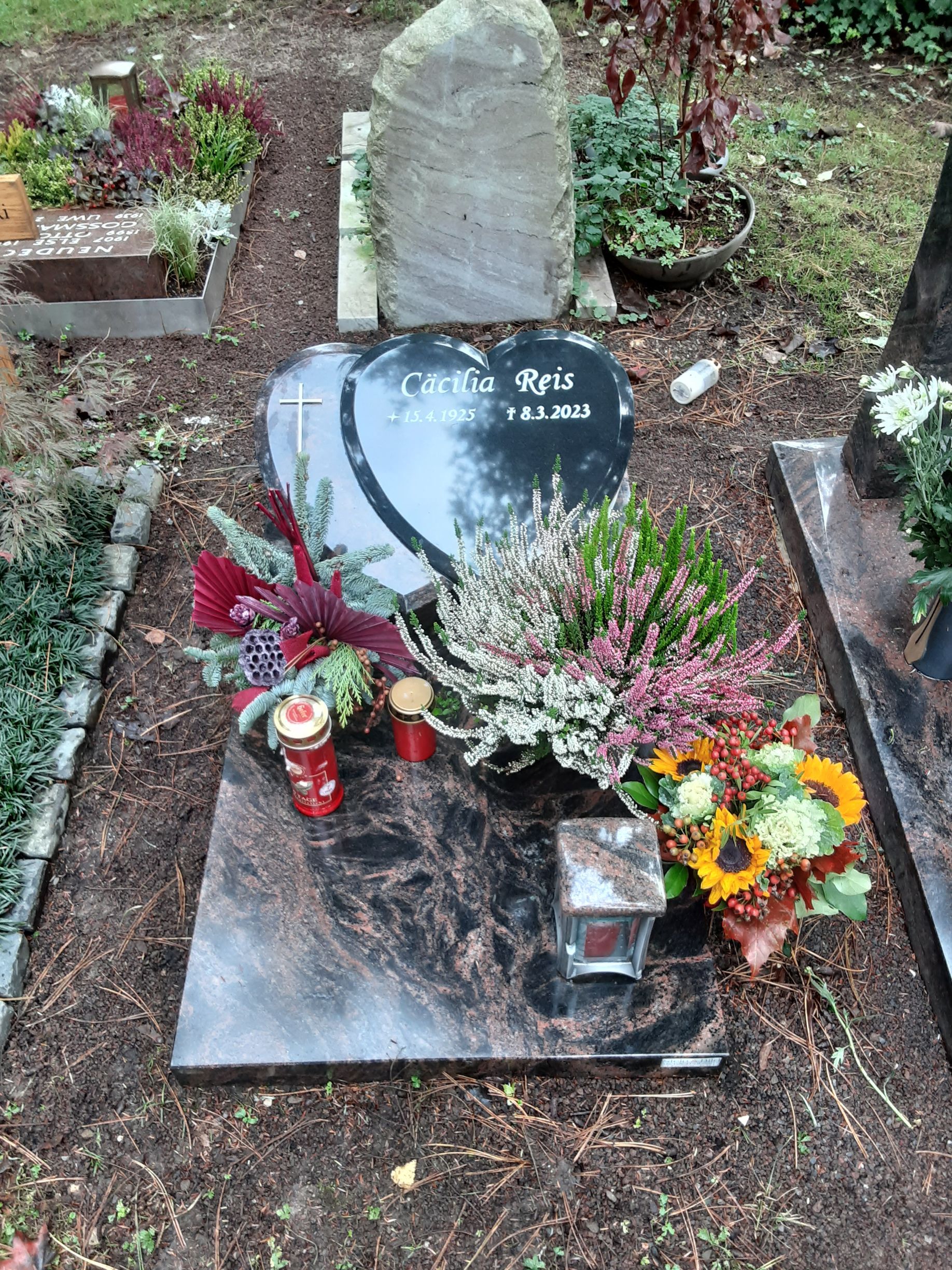 Grab mit schwarzem, herzförmigem Grabstein und Blumen. Kerze, Laterne und Steinmarkierung.