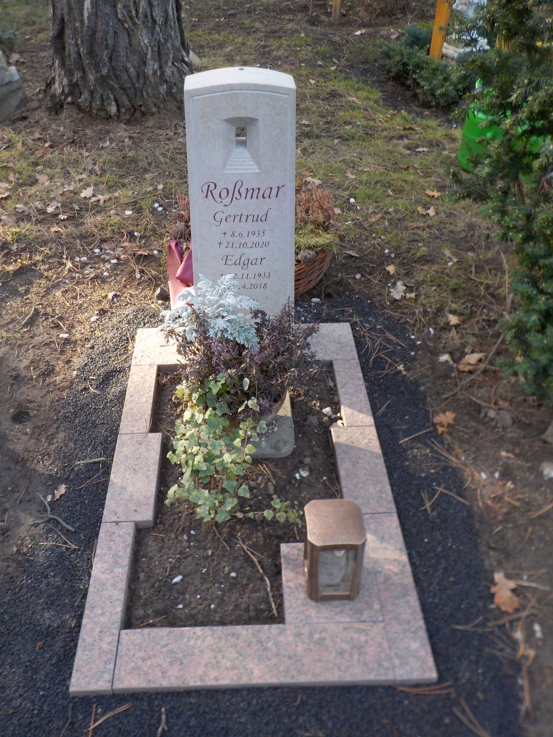 Ein rechteckiger Grabstein mit Glockenmotiv, umgeben von einem Rand und kleinen Pflanzen.