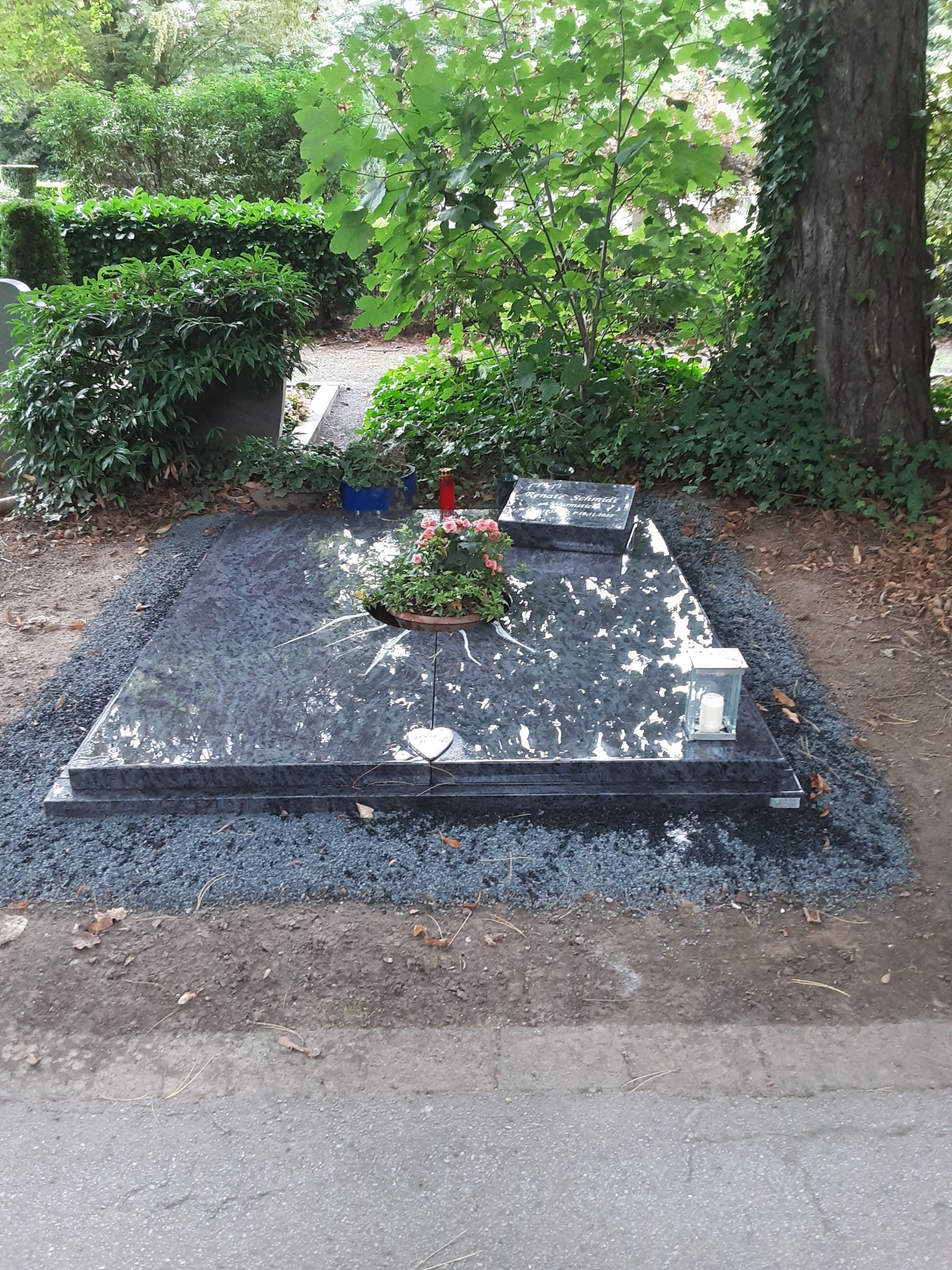 Ein dunkler Granitgrabstein mit Topfblumen auf einem Bett aus dunklem Kies. Im Hintergrund sind ein Baum und Grünpflanzen zu sehen.