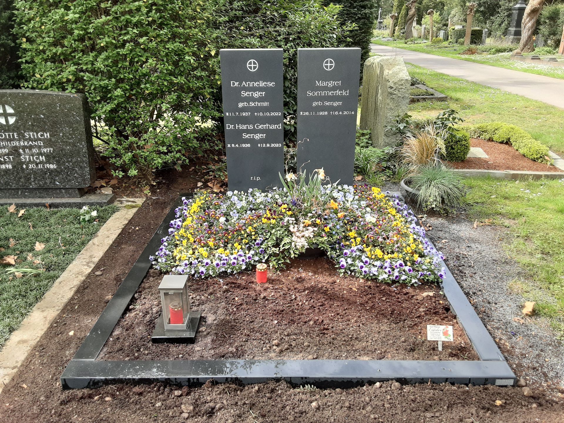 Grab mit zwei schwarzen Grabsteinen, Blumengesteck, einer Laterne und Einfassung auf einem Friedhofsgelände.