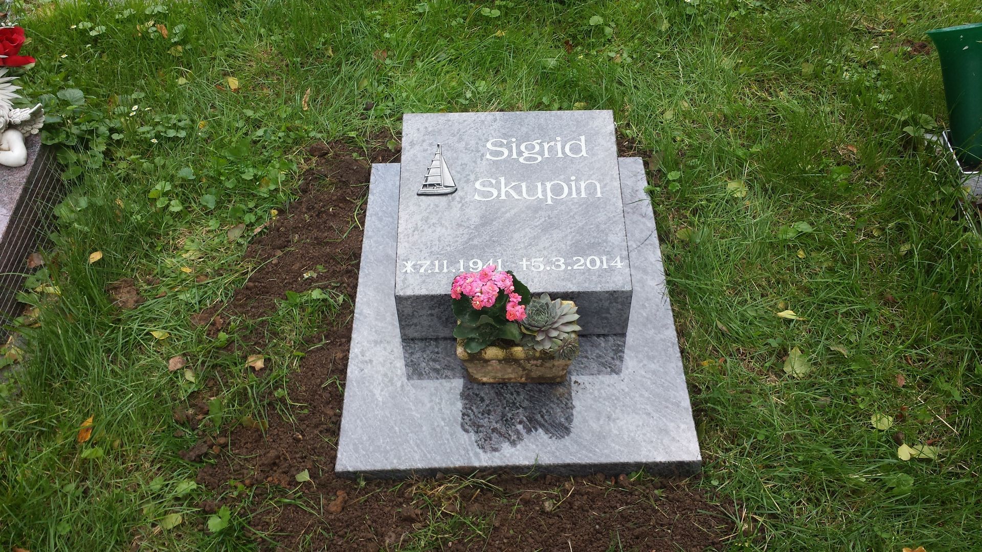 Grauer Grabstein mit rosa Blüten auf einer Rasenfläche. Name „Sigrid Skuolt“ und Lebensdaten sind in den Stein eingraviert.
