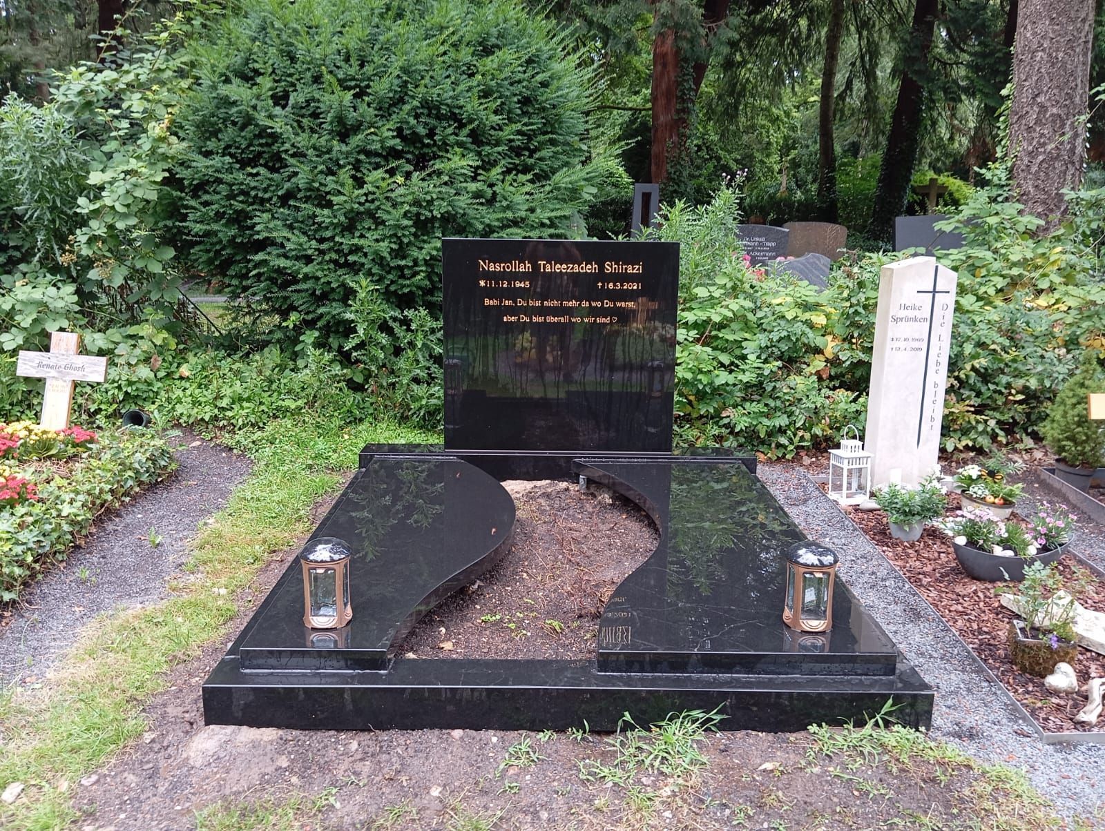 Schwarzer Granitgrabstein mit zwei gebogenen Abschnitten und einem vertikalen Grabstein auf einem Friedhof.