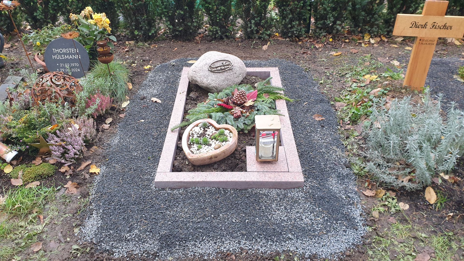 Grab mit steinernem Grabstein, herzförmigem Pflanzgefäß und Laterne, umgeben von dunklem Kies und Pflanzen.