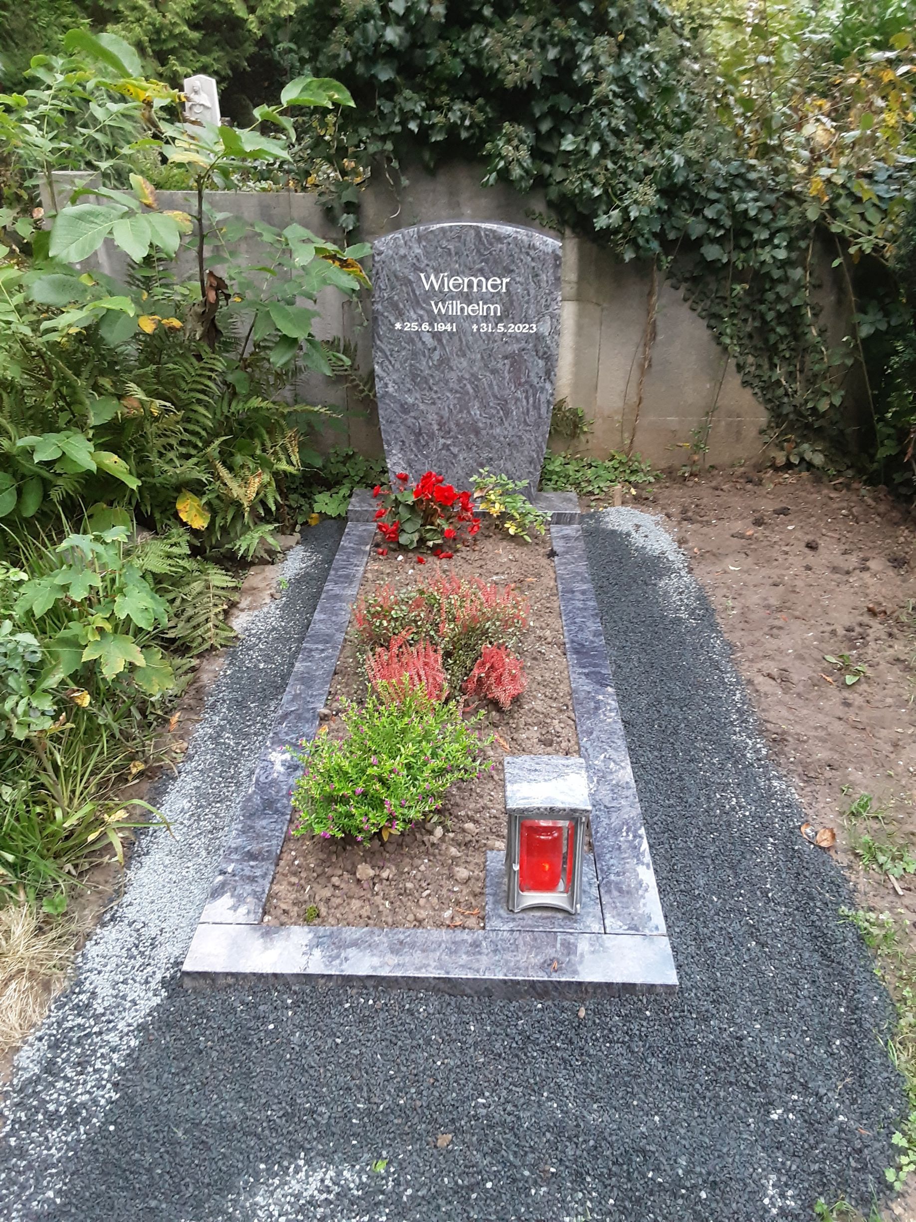Grabstein auf einem Friedhof mit Blumen, einer Laterne und dunkelgrauem Kies.