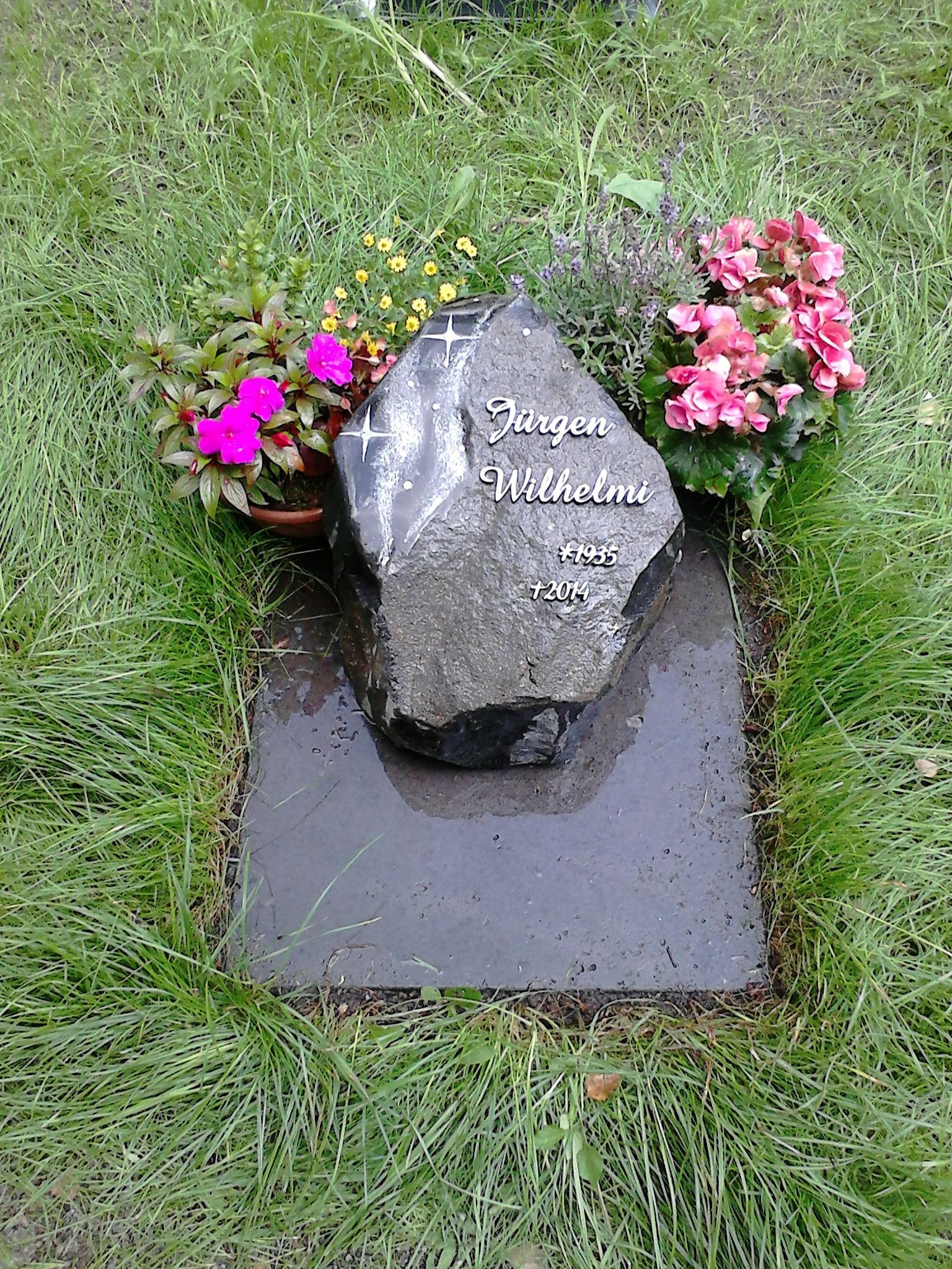 Grab mit einem dunklen, felsenförmigen Grabstein und bunten Blumen; eingebettet in Gras.