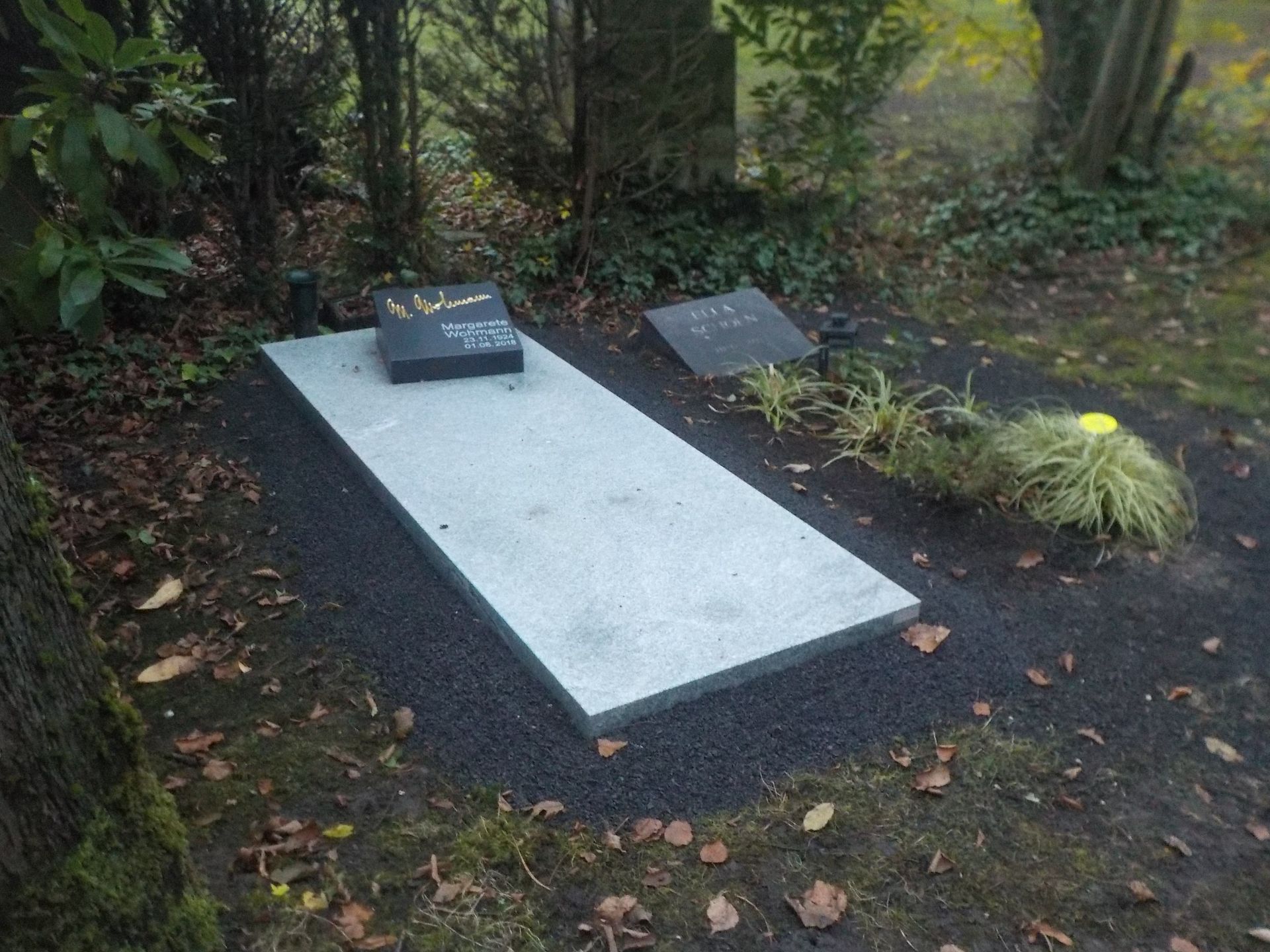 Ein rechteckiger Granitgrabstein auf einem Friedhof, daneben ein kleinerer Grabstein und ein kleines Stück Rasen.