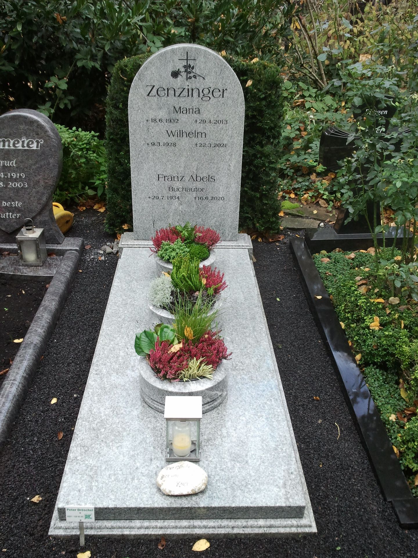 Grabstein mit drei Blumenarrangements, einer brennenden Kerze und einer dunklen Steineinfassung.