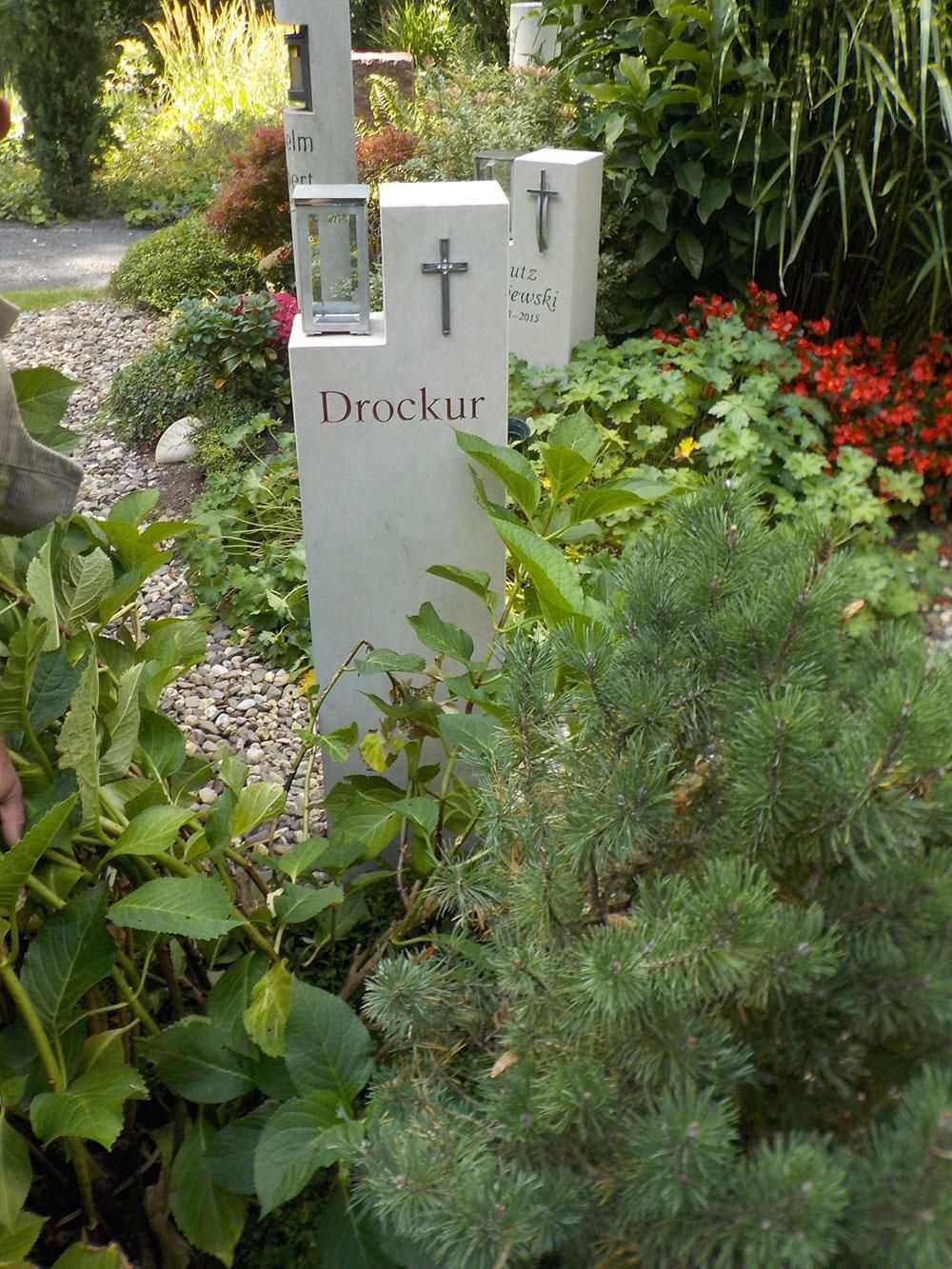 Ein Grabstein mit einem Kreuz darauf befindet sich in einem von Pflanzen umgebenen Garten.