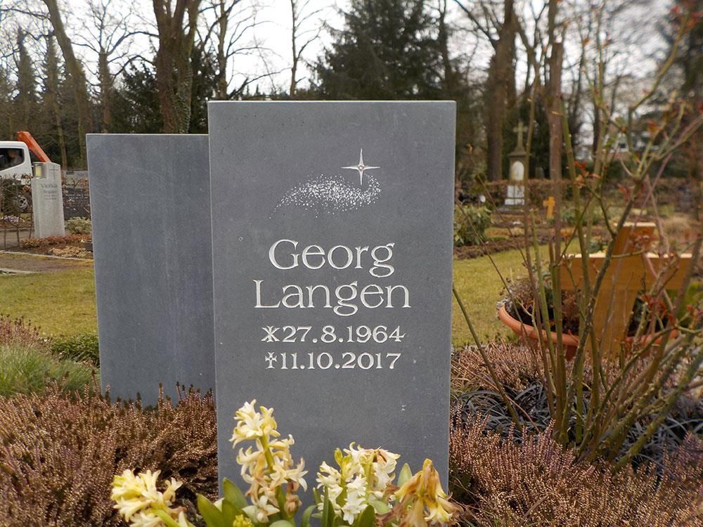 Ein Grabstein  steht auf einem von Blumen umgebenen Friedhof.