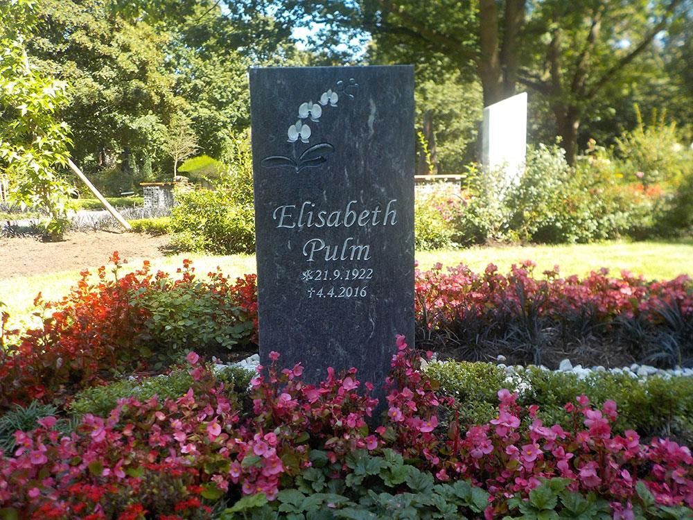 Ein Grabstein ist von rosa und roten Blumen umgeben.