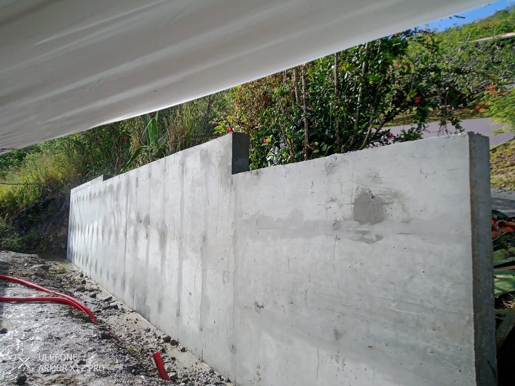 Un mur de soutènement en béton gris, nouvellement construit, se dresse à l'extérieur, adossé à une colline arborée et végétalisée.