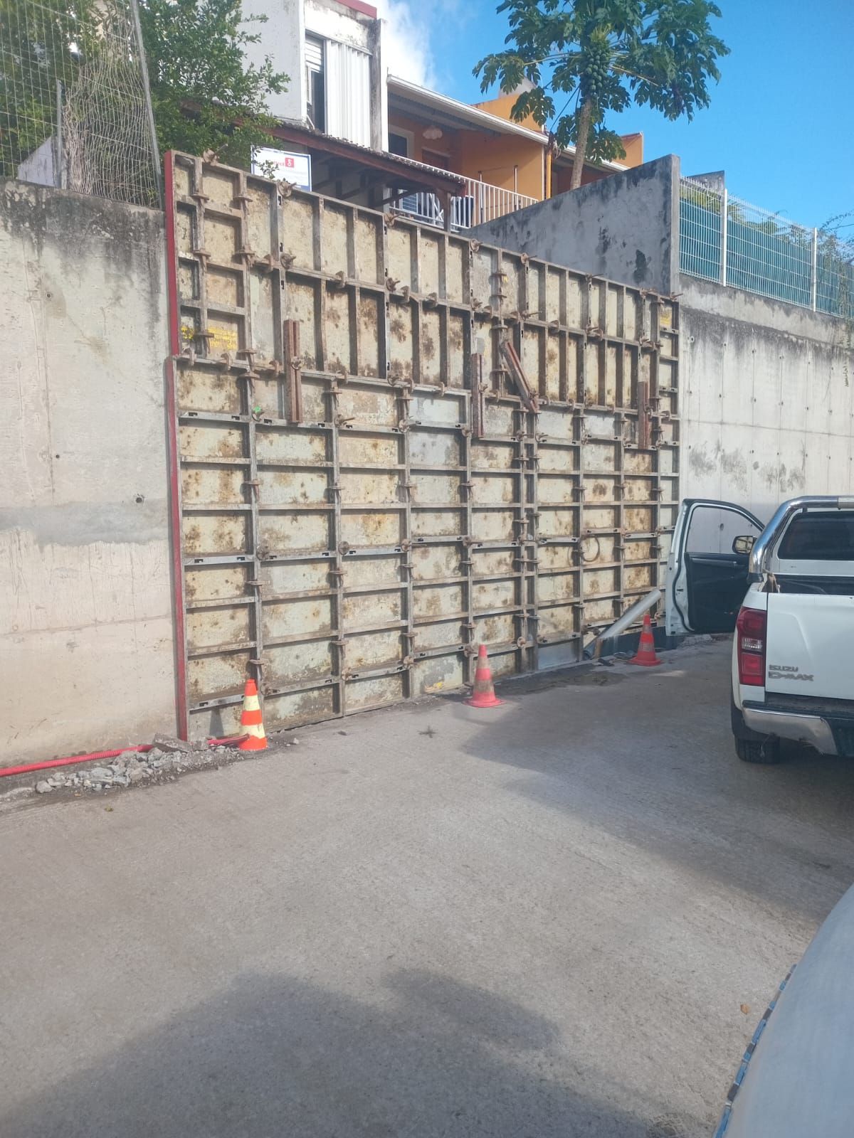 Des coffrages modulaires en métal pour béton sont installés contre un mur en béton existant dans une aire de stationnement délimitée par des cônes de signalisation.