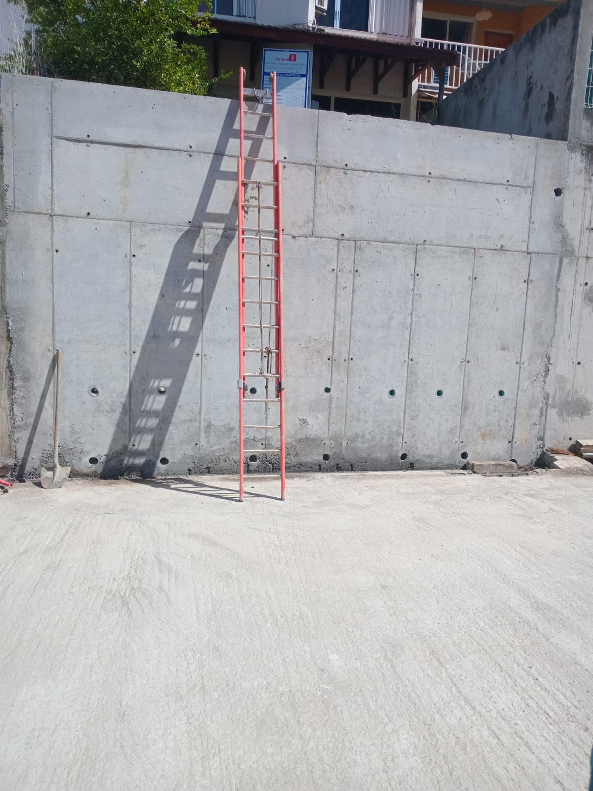 Une échelle orange est appuyée contre un haut mur de béton gris clair, en extérieur, sous un soleil éclatant.