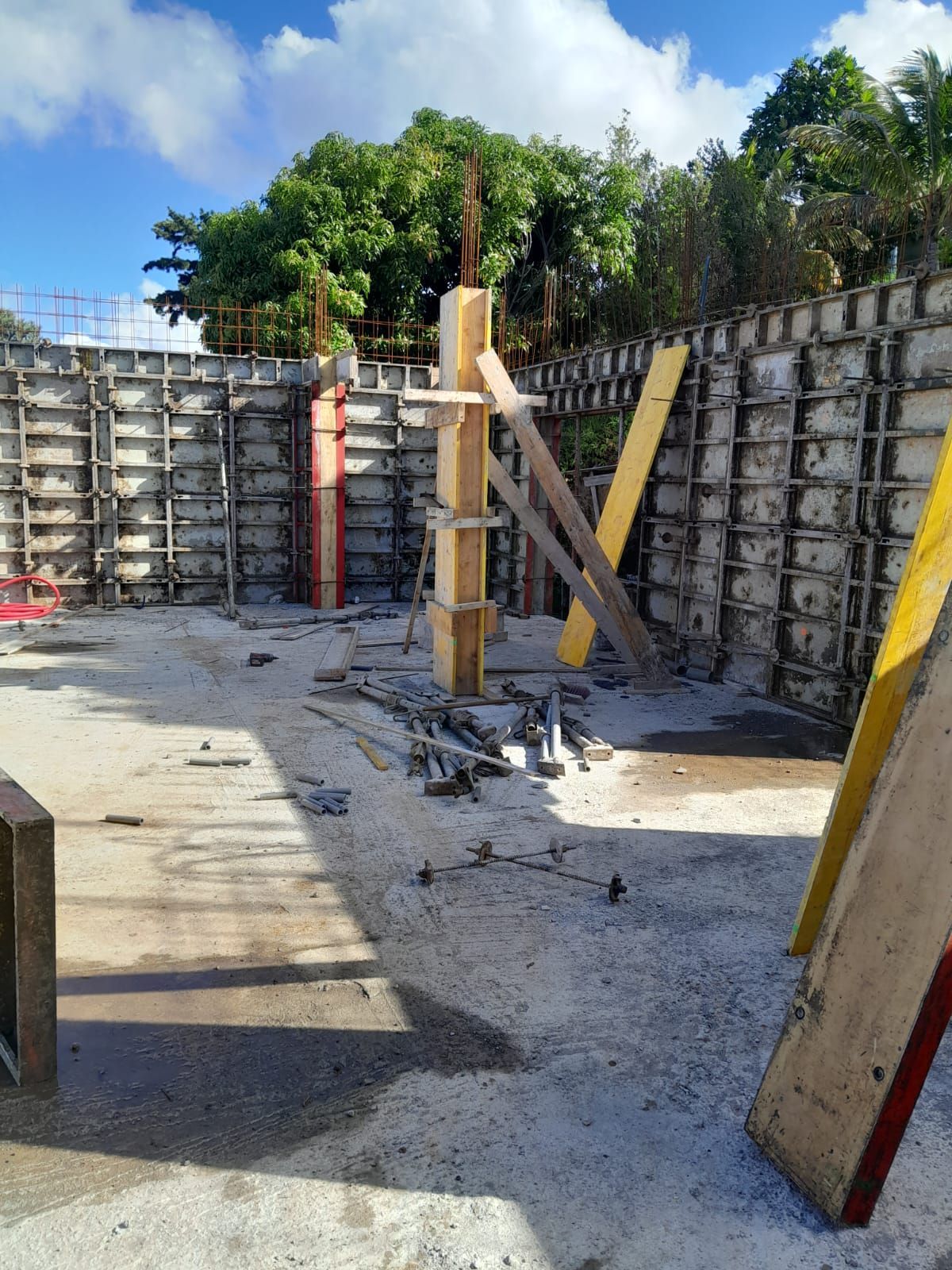 Chantier de construction avec des murs en coffrage métallique et un pilier de soutien en bois central, sous un ciel d'un bleu éclatant.