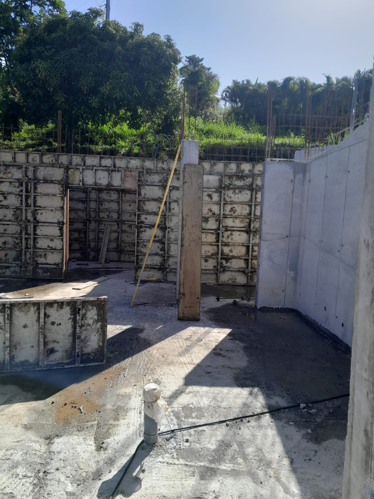 Un chantier de construction avec des murs de fondation en béton, des coffrages de soutènement et une canalisation verticale sous un ciel bleu azur.