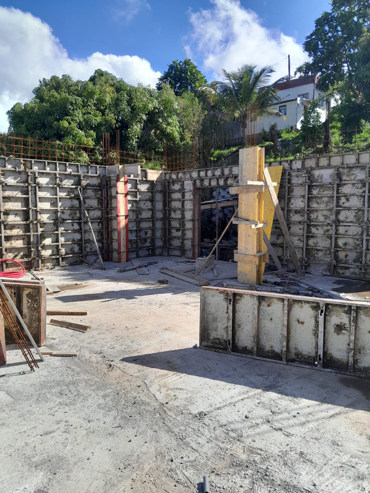 Chantier de construction montrant des murs en béton avec des coffrages à ossature métallique, un coffrage de colonne jaune vertical et un sol en terre battue nivelé.