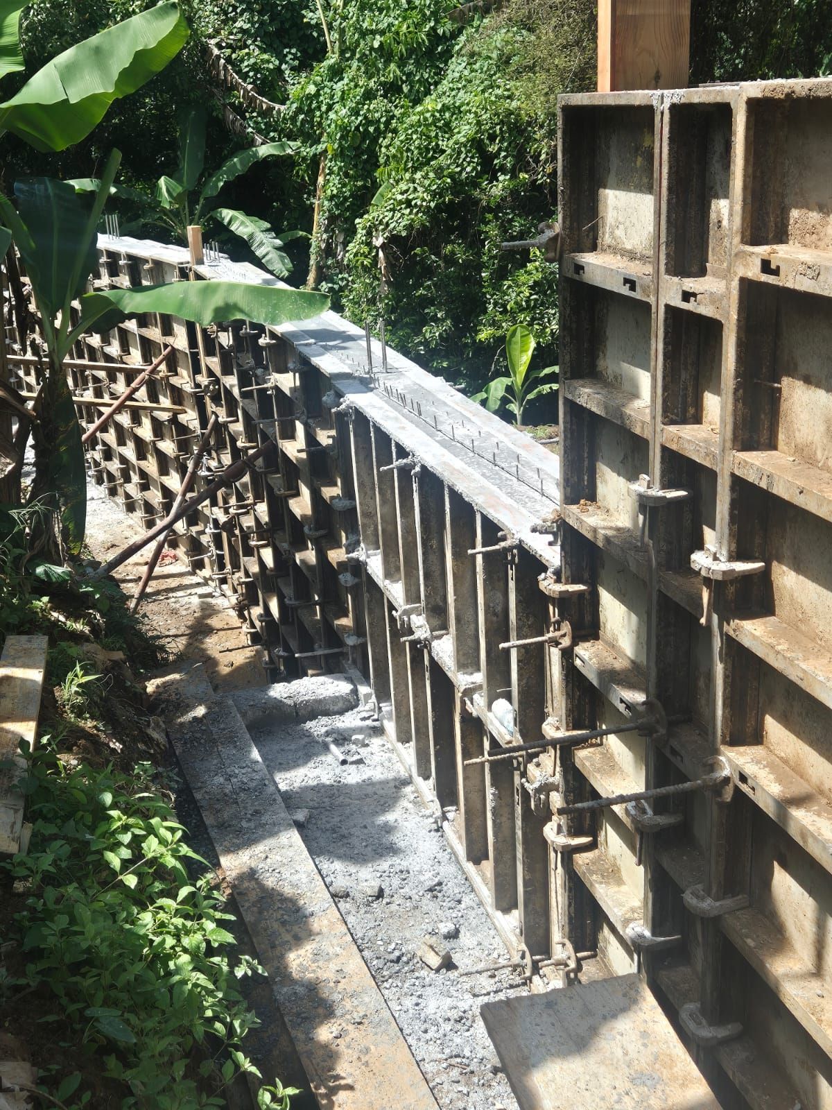 Coffrages de murs en béton installés sur un chantier de construction, avec une base de fondation en gravier dans un cadre extérieur verdoyant et luxuriant.