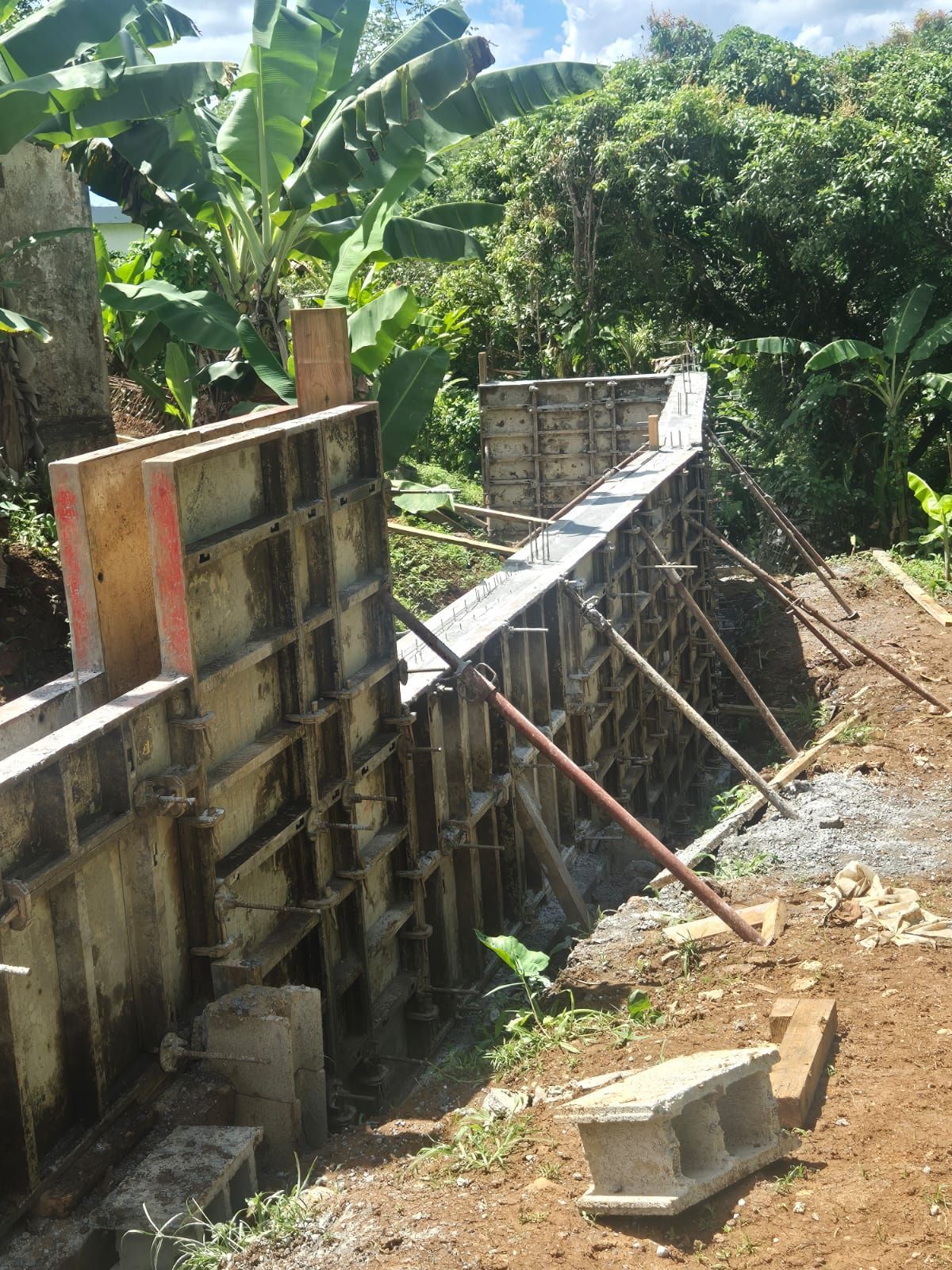 Panneaux de coffrage en acier pour un mur de soutènement en béton en construction sur un talus de terre avec une végétation luxuriante en arrière-plan.