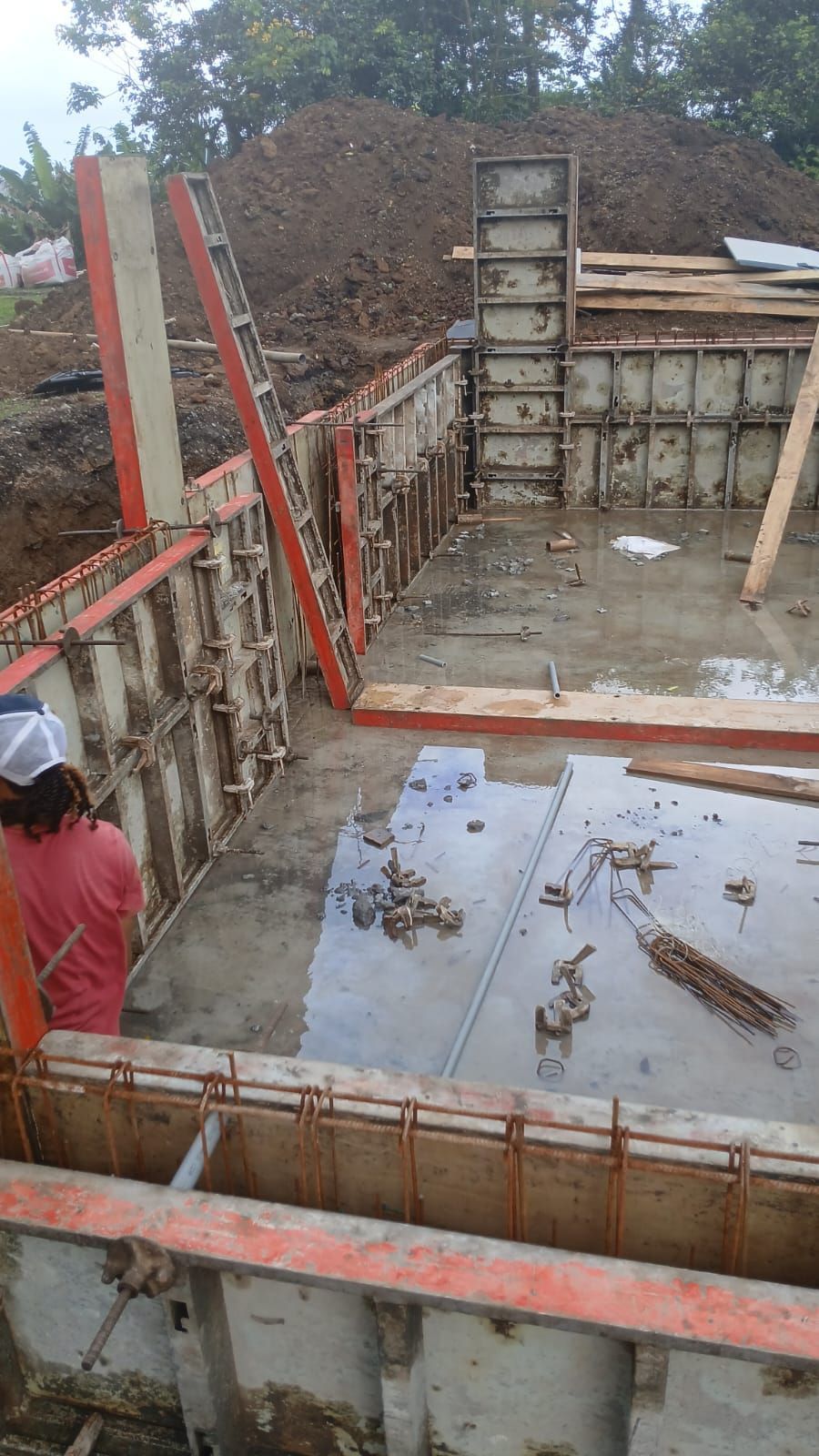 Sur un chantier de construction, une personne vêtue d'une chemise rouge travaille à l'intérieur de coffrages en bois et en métal destinés à accueillir des fondations en béton coulé.