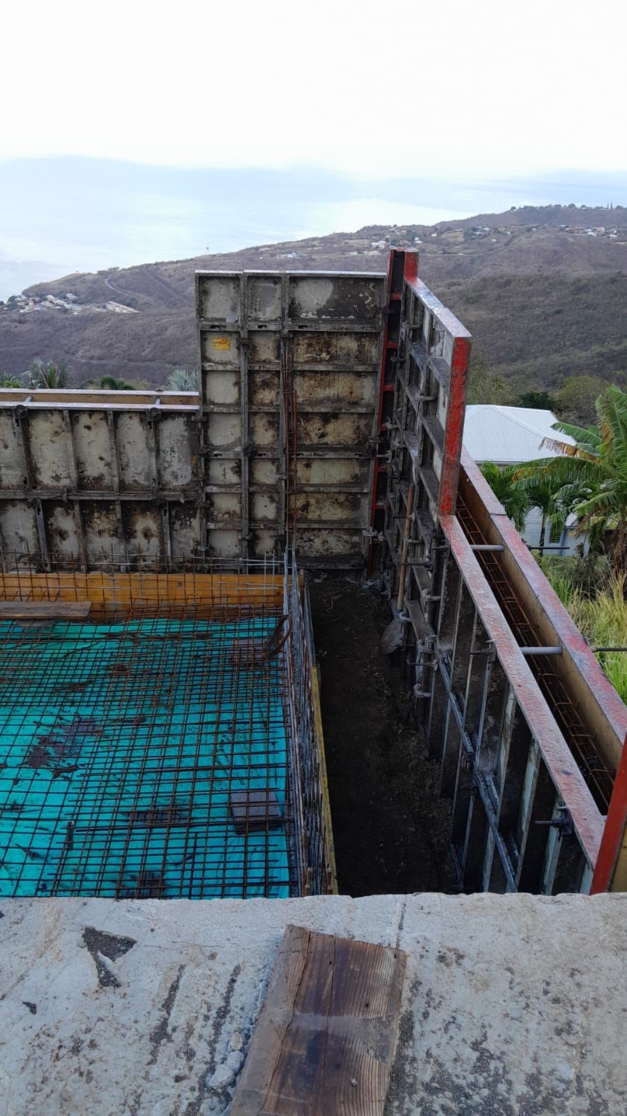 Chantier de construction montrant des coffrages métalliques en béton installés sur une armature en fer à béton, avec une vue sur une colline en arrière-plan.